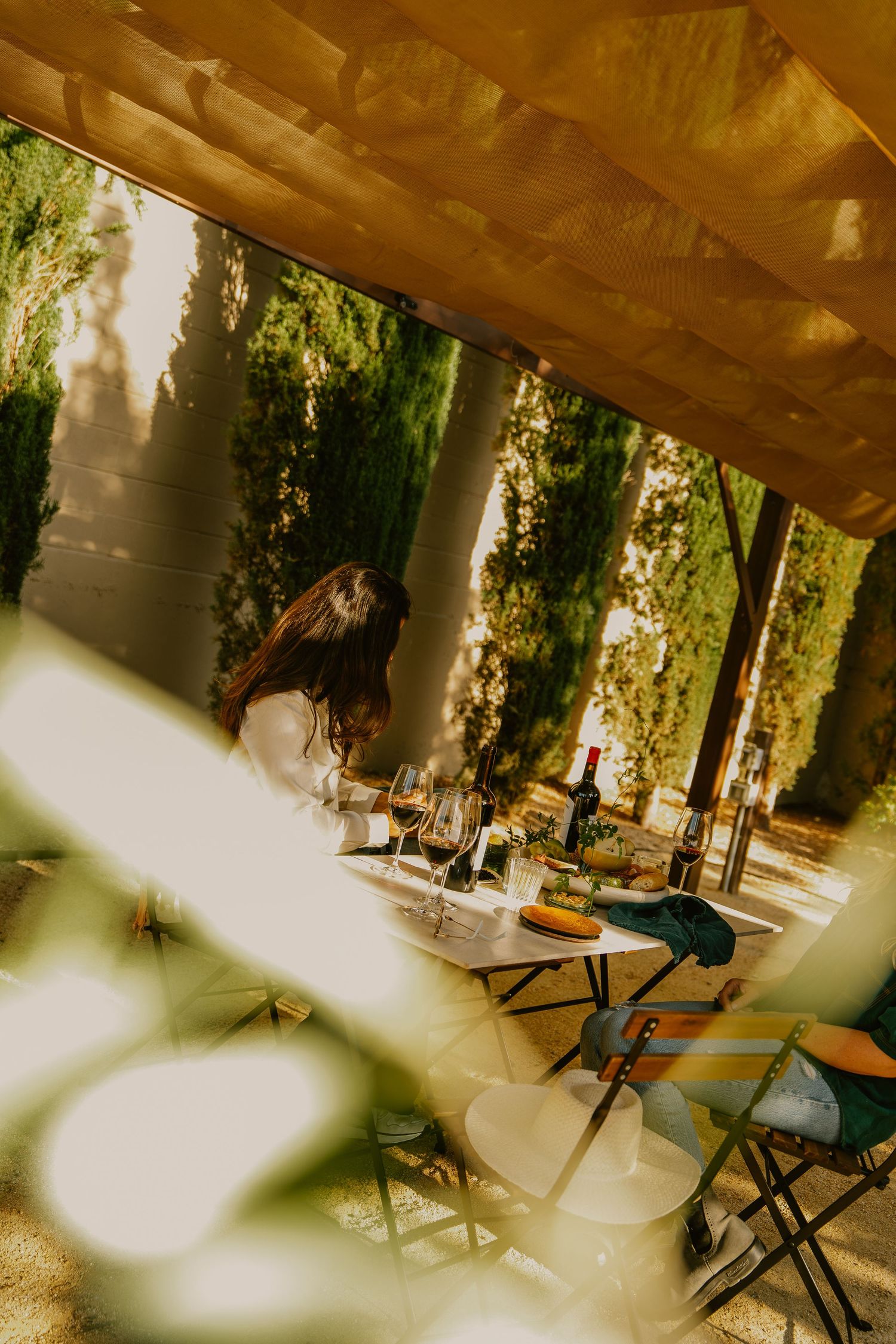 Woman enjoying wine tasting at outdoor table surrounded by tall cypress trees and golden light.