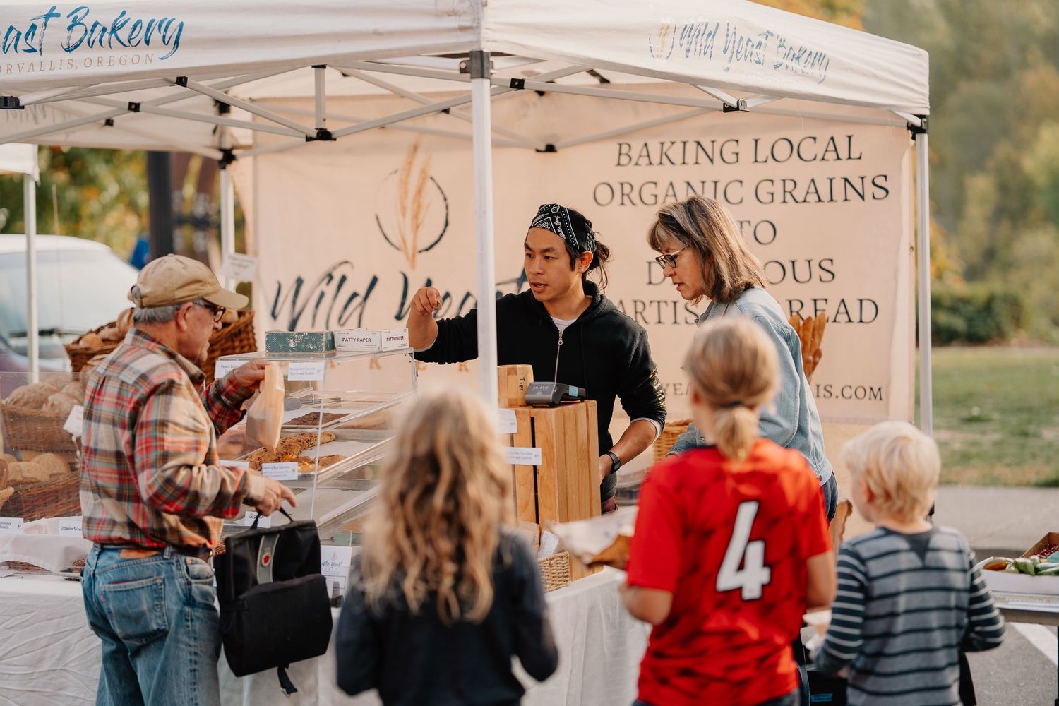 Local bakers at farmers market tent display fresh bread and baked goods to customers on sunny afternoon.