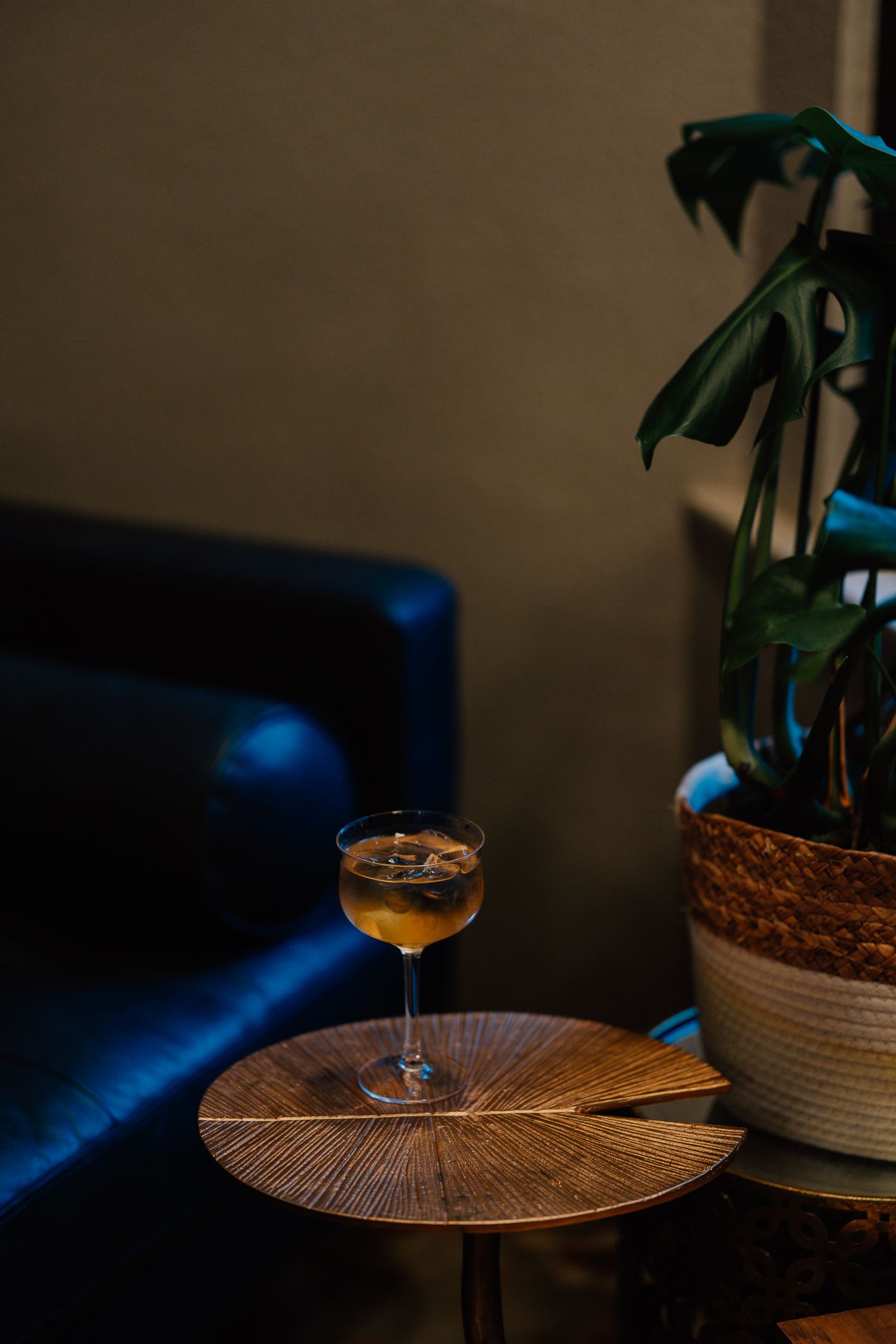 An amber cocktail in a coupe glass sits on a round wooden table next to a potted plant in a dark moody setting.
