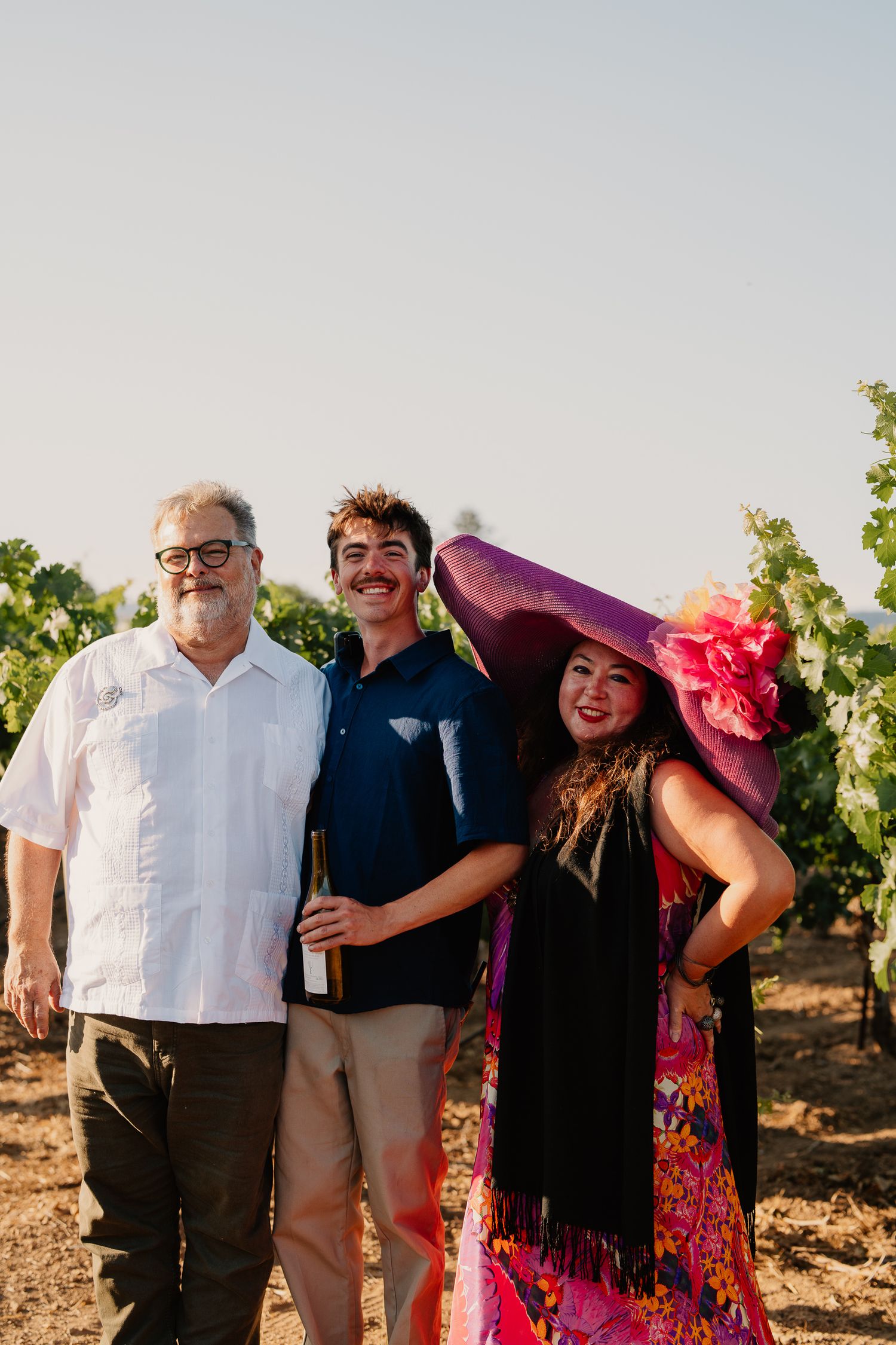 Three people pose together in a vineyard wearing colorful outfits during a wine tasting event.