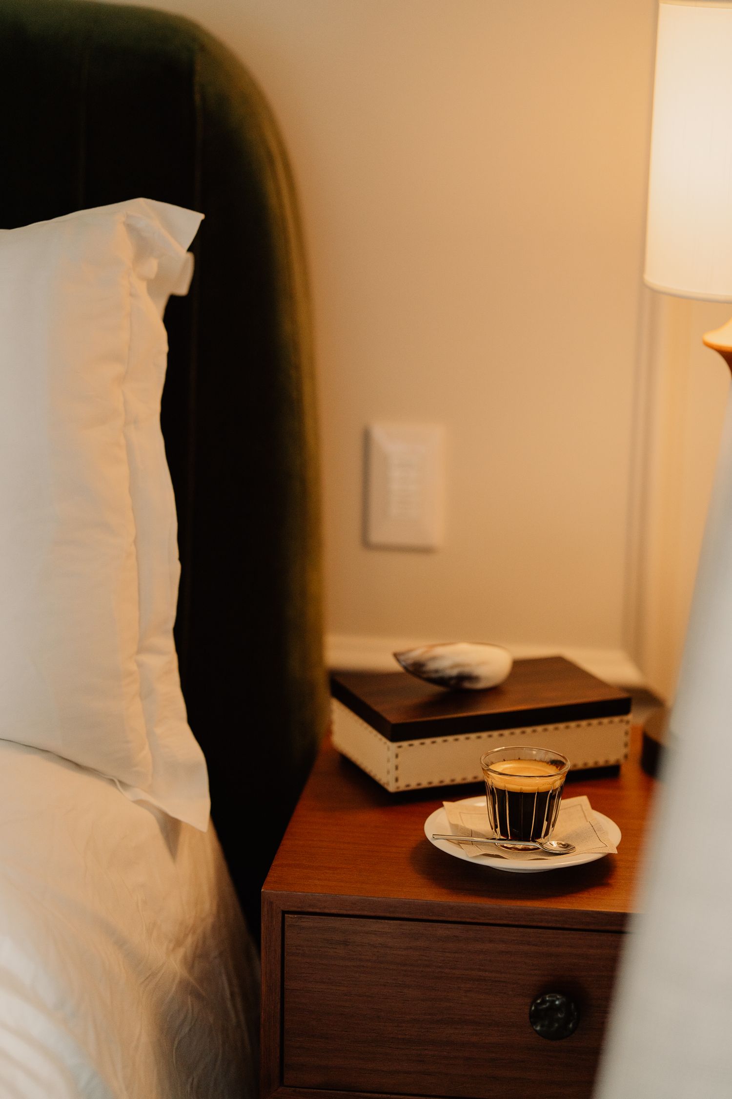 A hotel bedside table with a white lamp, pillows, and a small plate with a chocolate dessert.