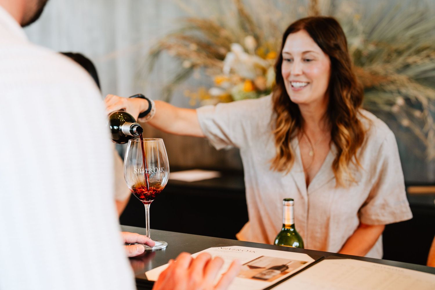 A smiling woman pours red wine into a glass at a winery tasting bar with a white wine bottle nearby.