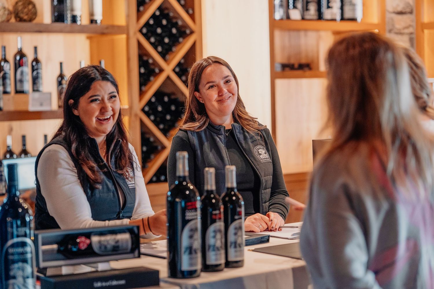 Two smiling women assist a customer at a winery tasting room counter with wine bottles displayed.