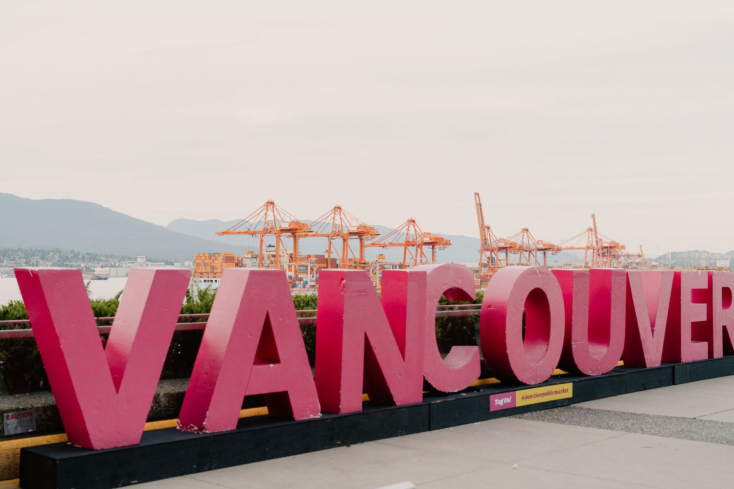 Giant pink letters spell out VANCOUVER against a scenic waterfront view with port cranes and mountains in the background.
