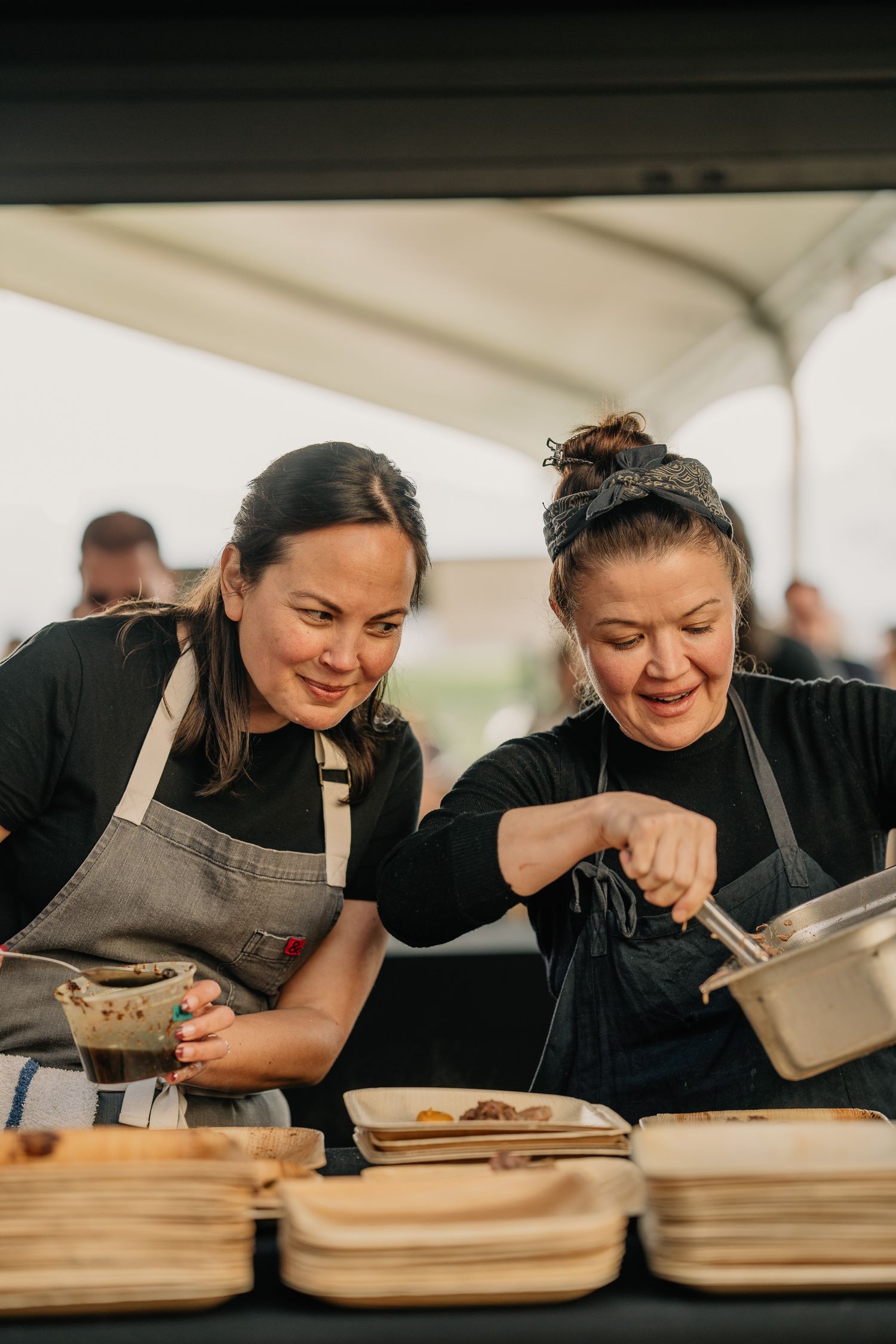 Two female chefs in black aprons collaborate while plating dishes at an outdoor food festival.