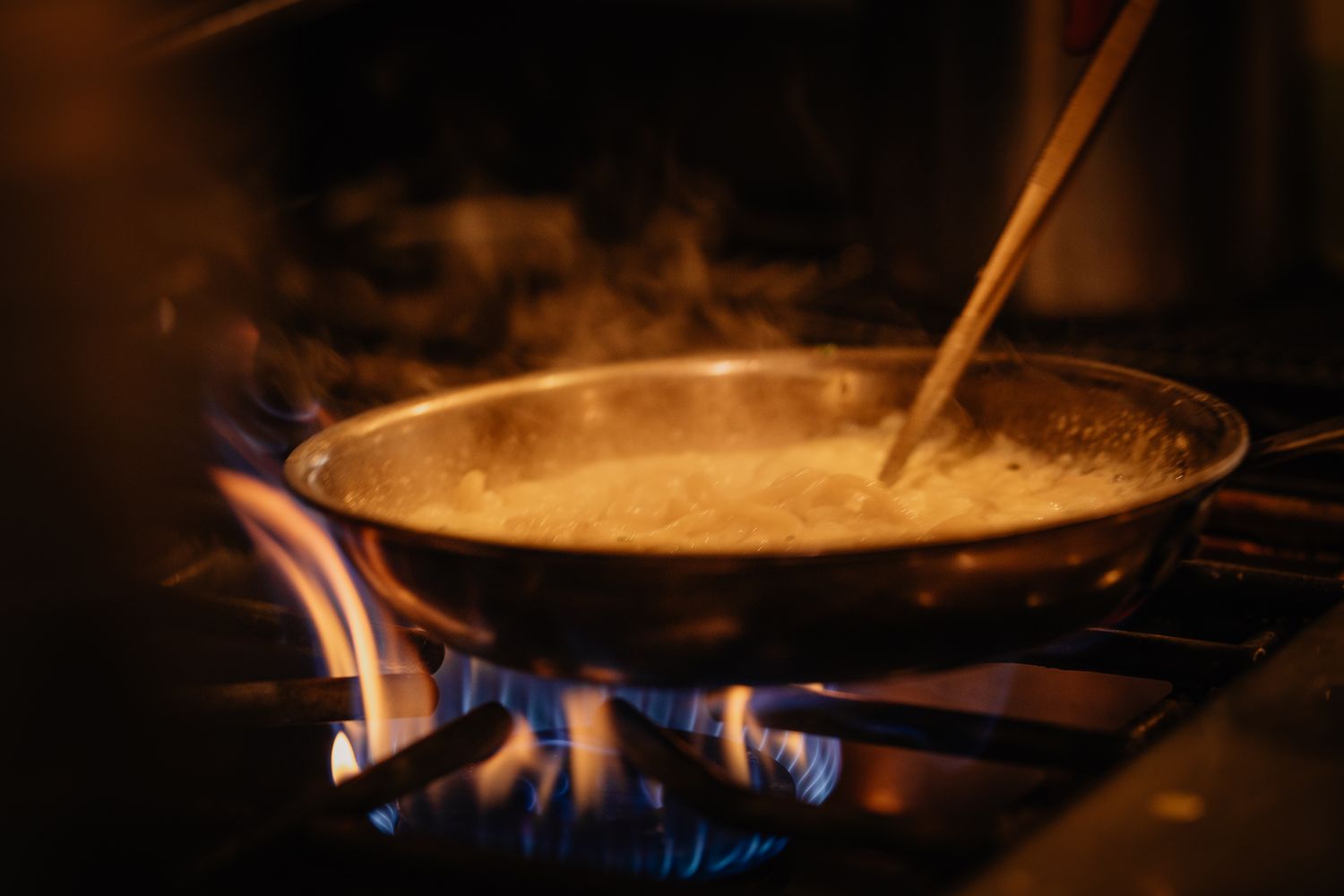 Golden sauce simmering in a copper pan over blue gas flame with steam rising in a dark kitchen.