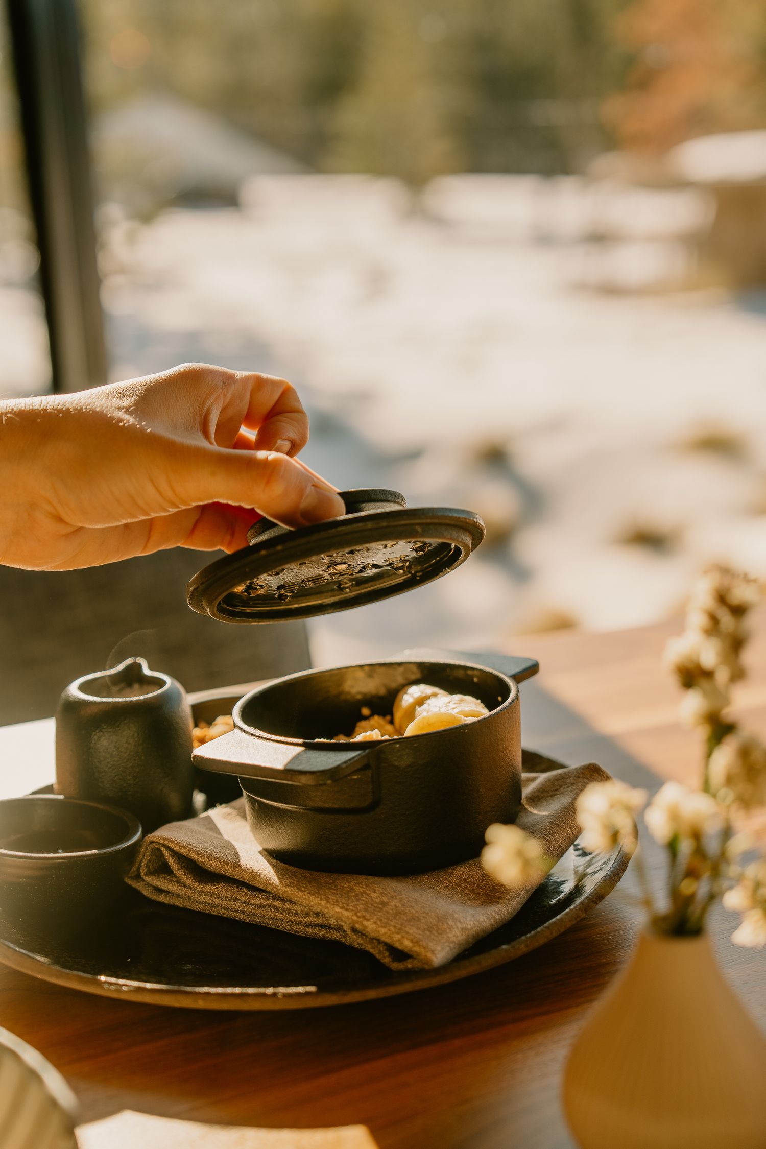 A hand lifts the lid of a cast iron pan to reveal a steaming dish in warm lighting.