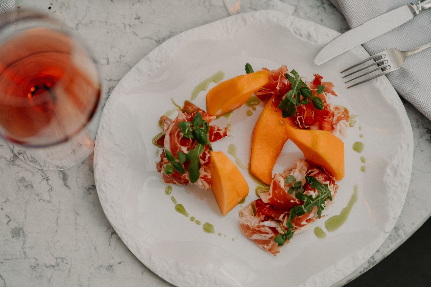 Elegant plate of cantaloupe melon with prosciutto and arugula, drizzled with herb oil, served alongside a glass of rosé wine.