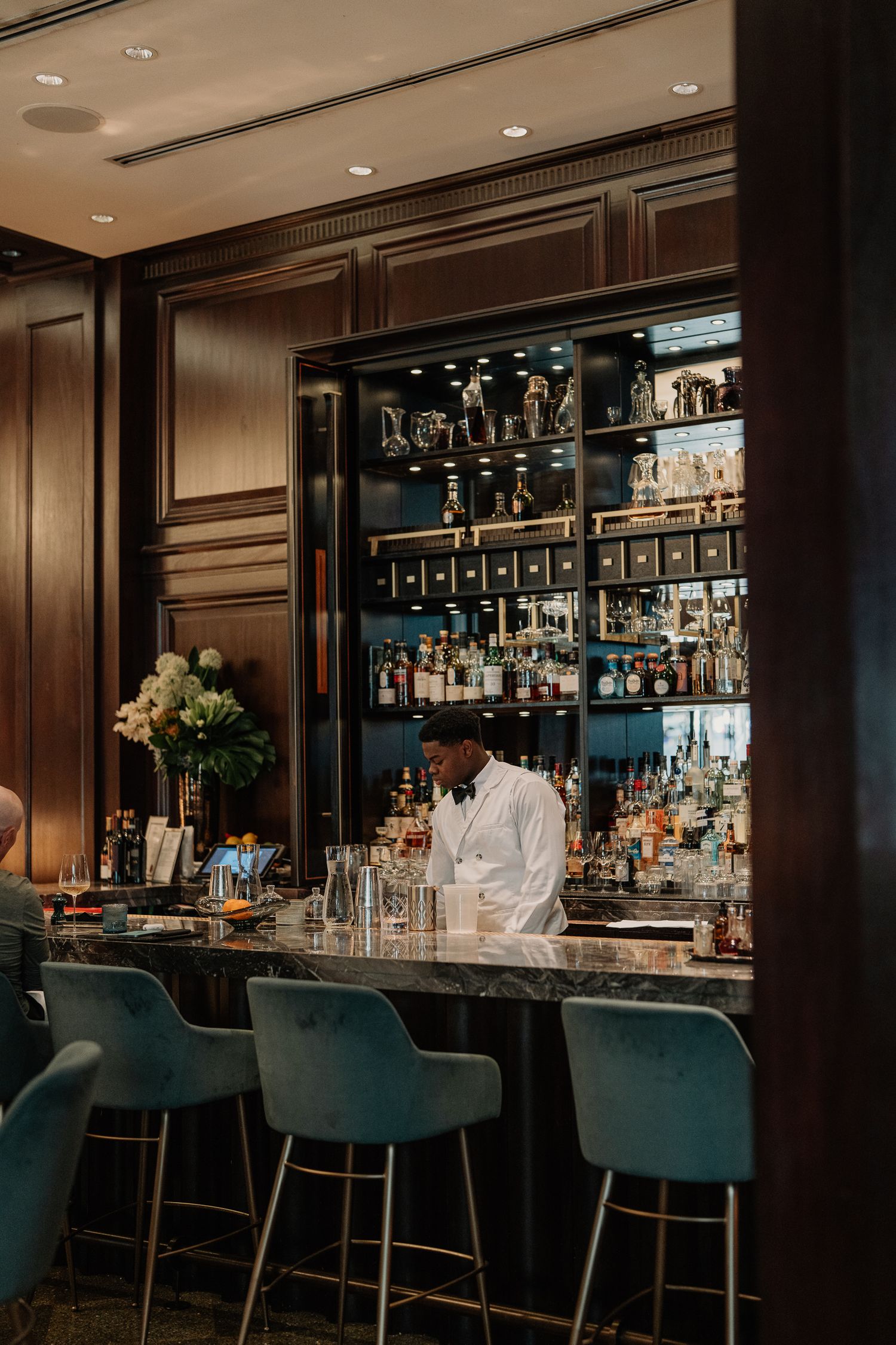 An elegant bar interior with wood paneling features teal velvet barstools and illuminated liquor displays.