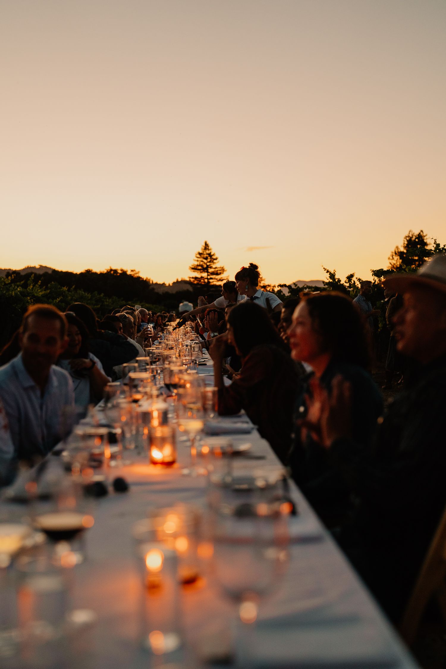 Long white dining table set for an outdoor dinner party at sunset with glowing candle centerpieces casting warm light.