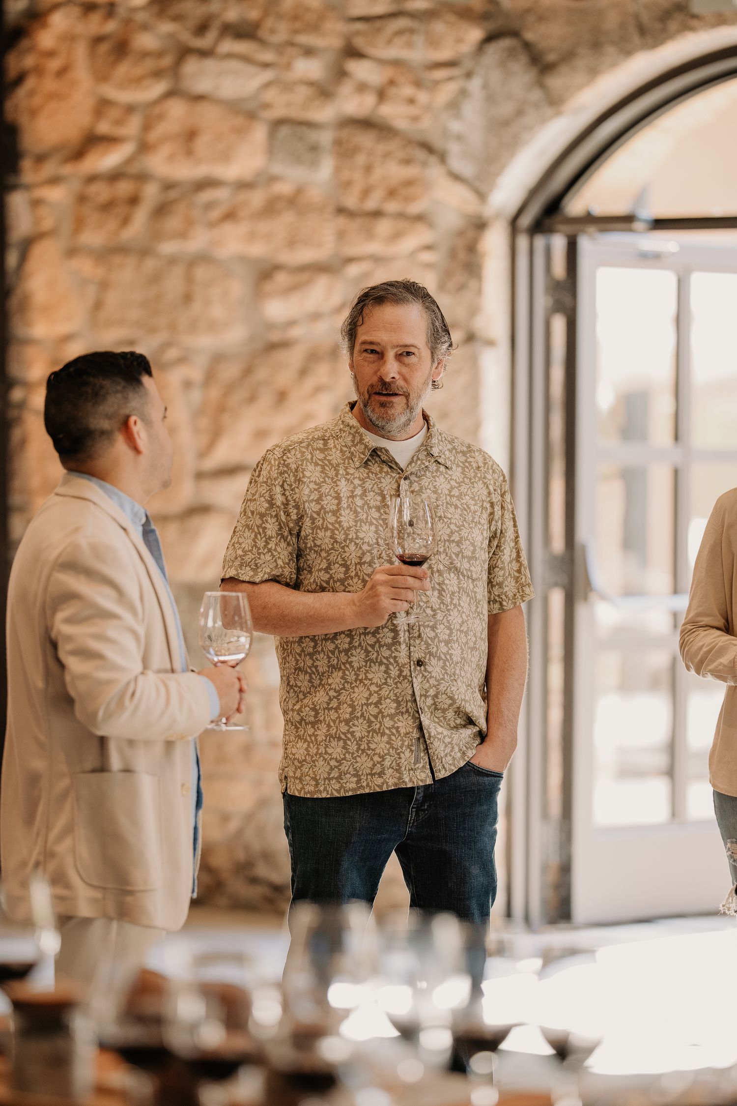 Two men enjoying wine tasting at a rustic stone winery, engaged in conversation while holding glasses of red and white wine.