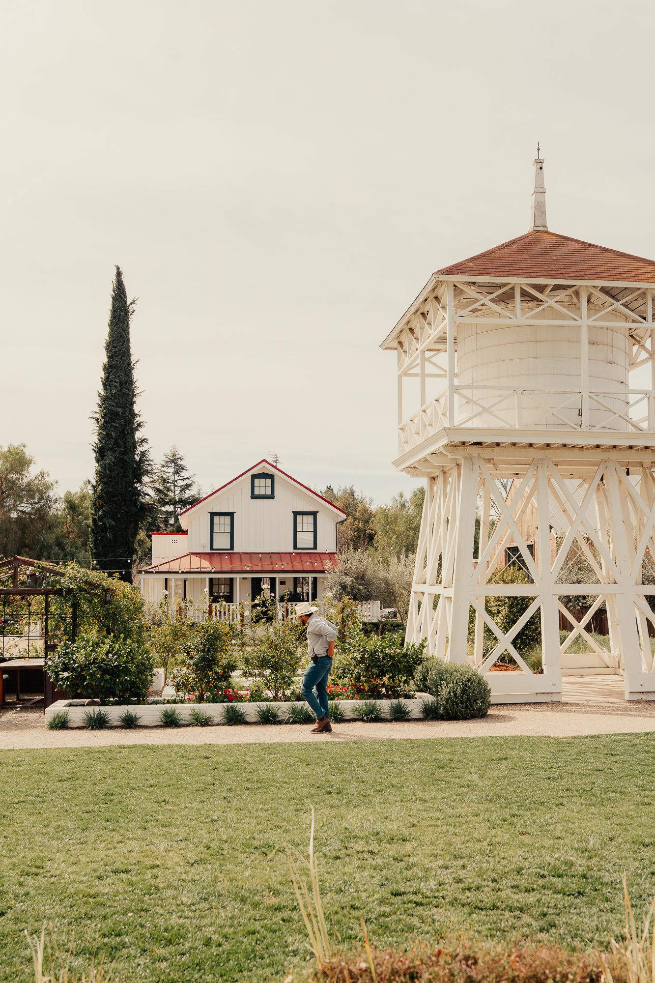 Person walking past a historic white water tower and farmhouse surrounded by lush gardens.