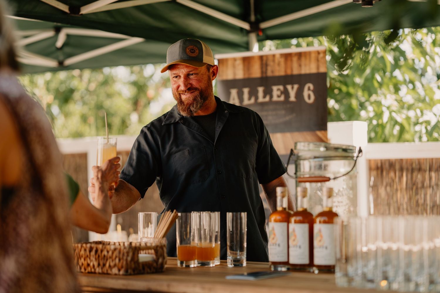 A bartender pours a signature cocktail at an outdoor bar counter in warm, natural lighting.