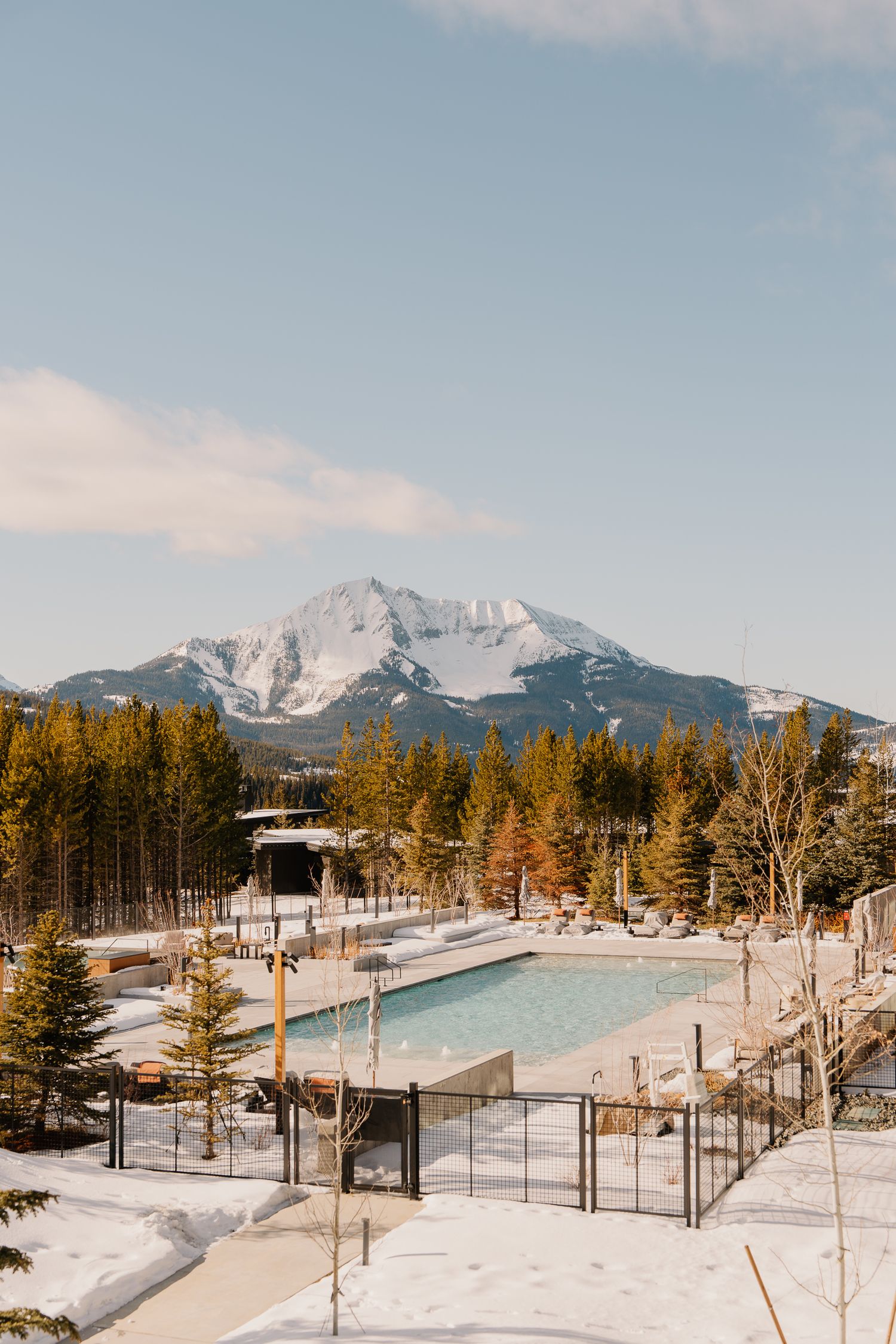 Snow-covered mountain resort with outdoor heated pool surrounded by evergreen trees in winter landscape.