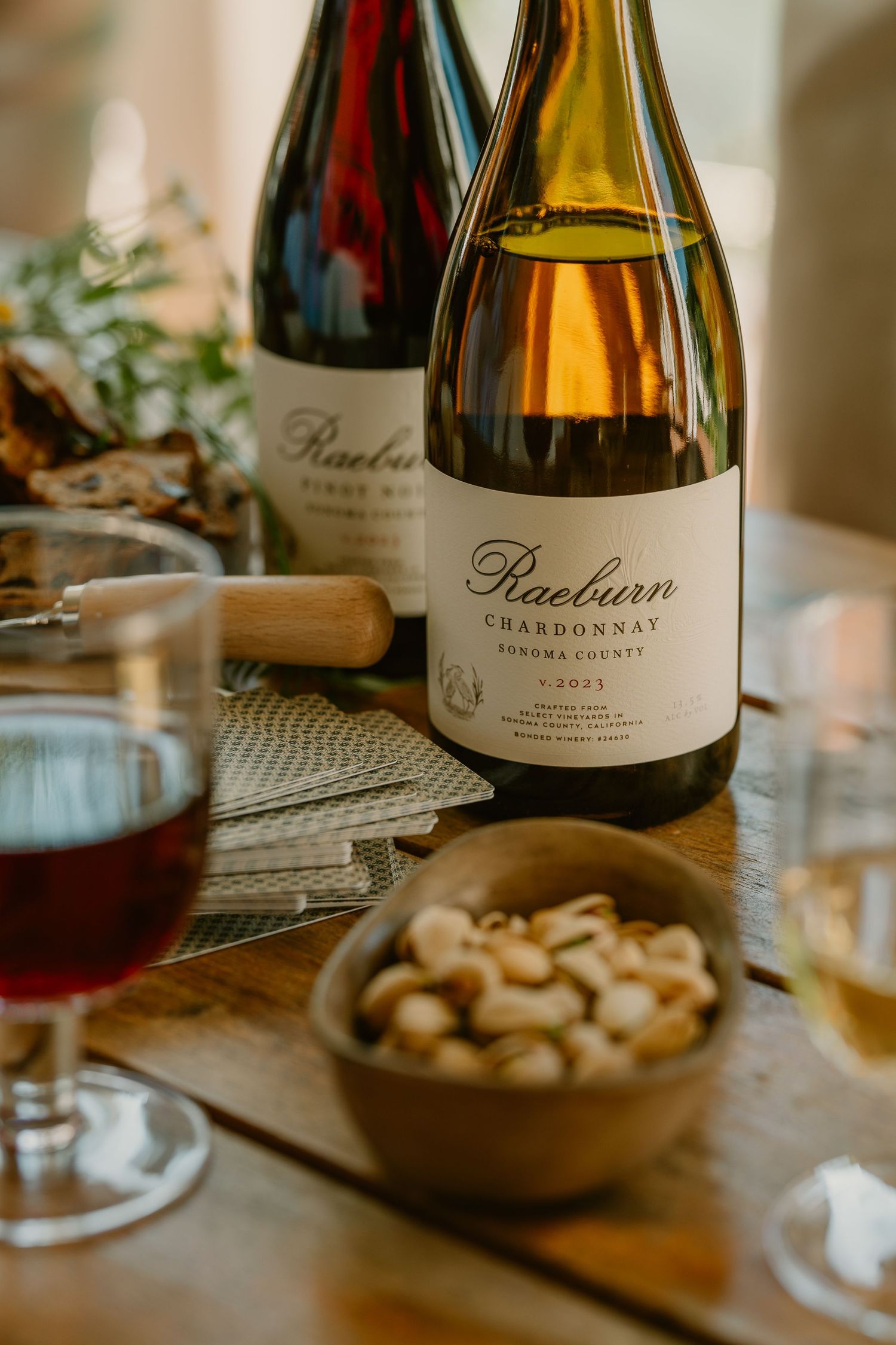 Raeburn Chardonnay Sonoma County wine bottle on rustic table with red wine glass and bowl of nuts.