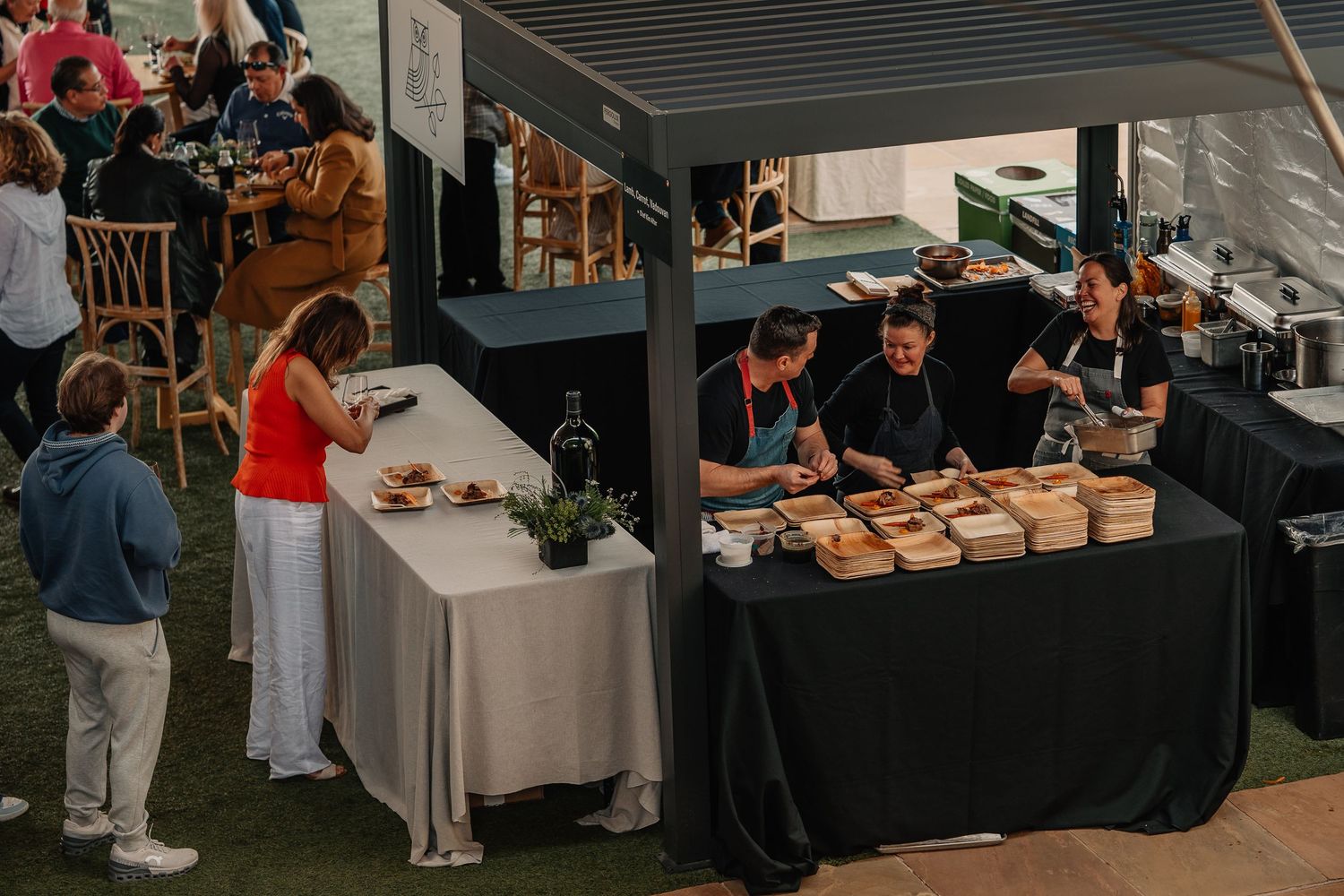 Chefs serving food samples at an outdoor culinary event with guests browsing wooden box displays at a black draped booth.