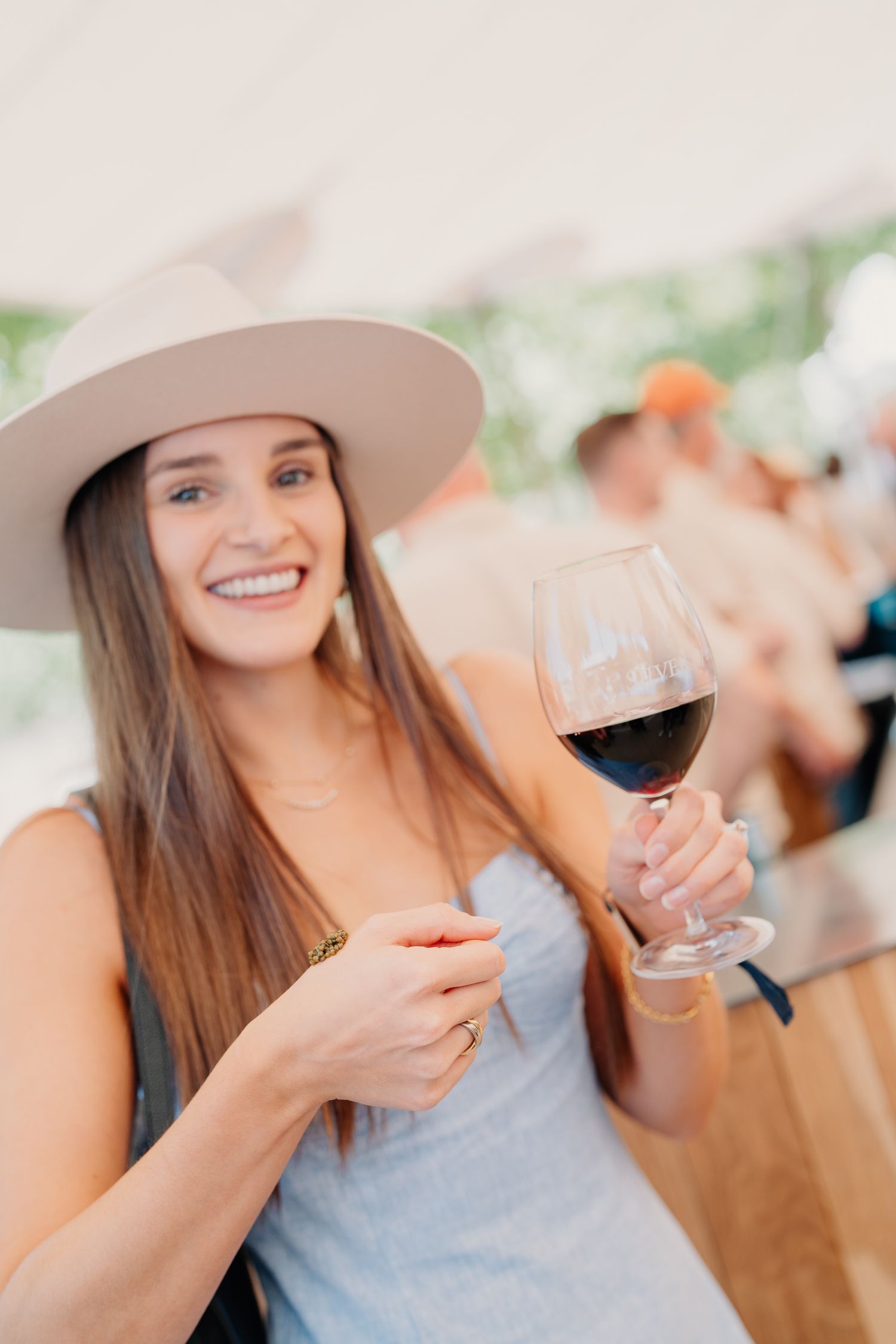 Smiling brunette woman in wide-brim hat holding red wine glass at outdoor winery tasting event.