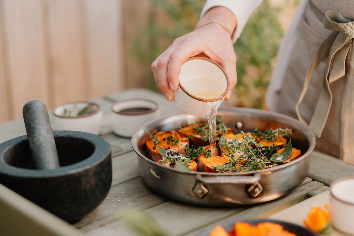 Chef pouring sauce over colorful roasted vegetables in a copper pan on a rustic wooden table.