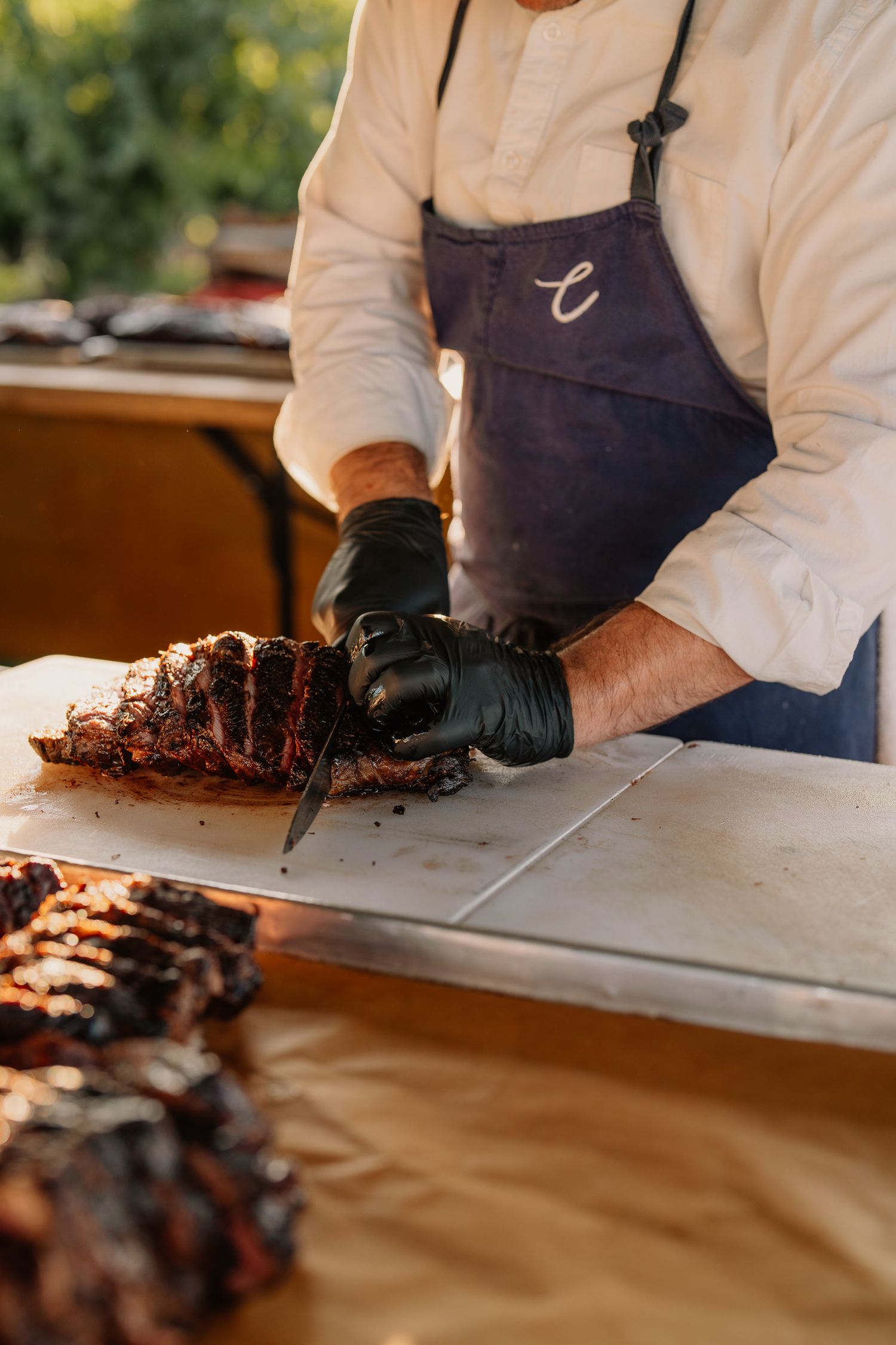Chef in navy apron and black gloves carving grilled meat on outdoor prep station.