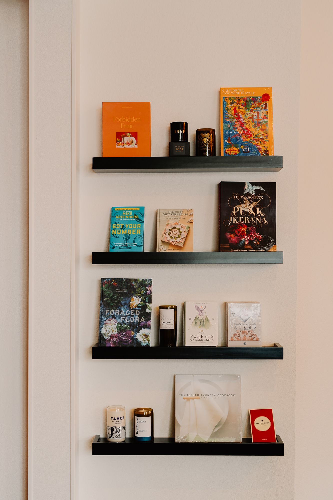 Four black floating shelves display an eclectic collection of books, candles, and decorative items against a white wall.