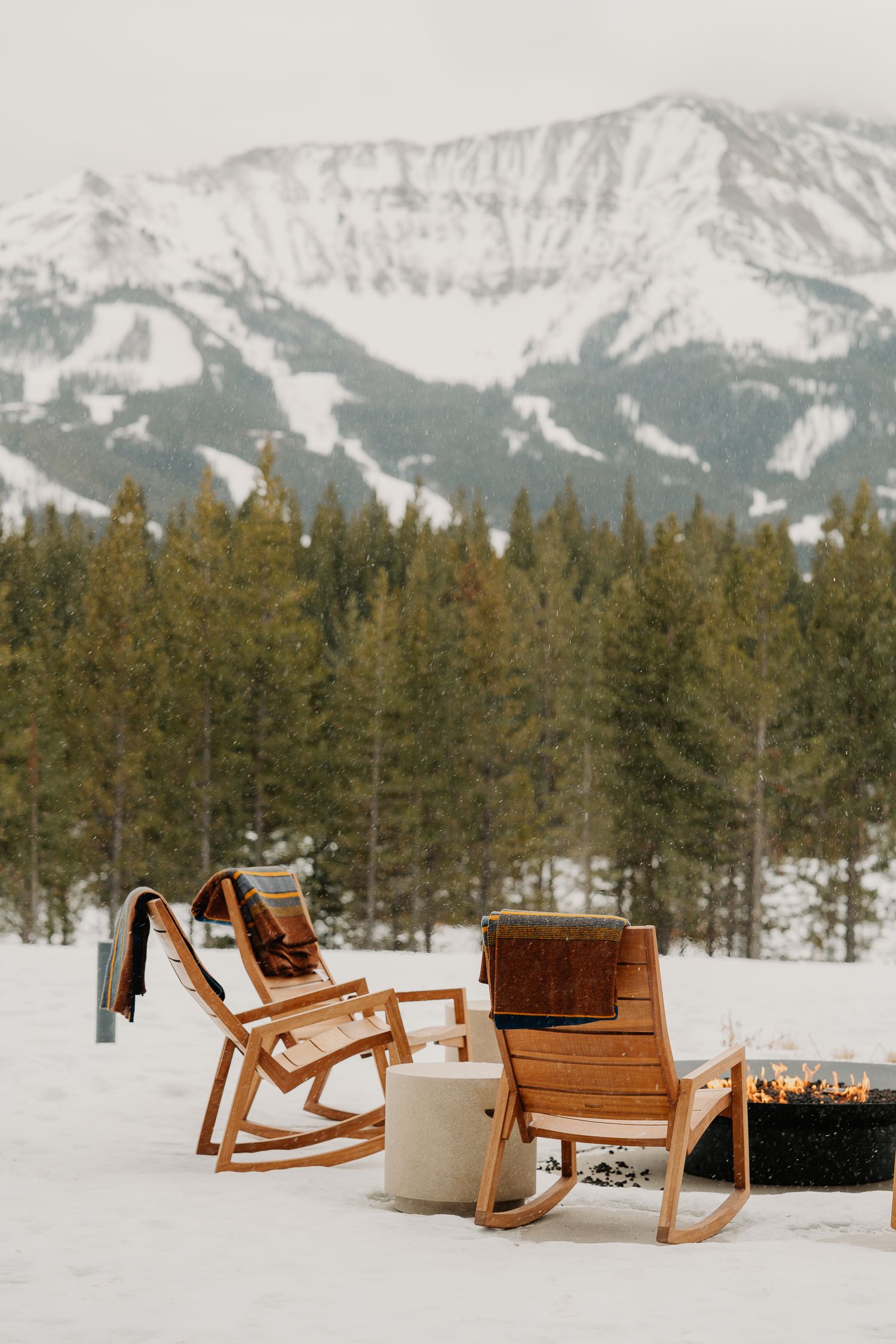 Wooden rocking chairs in snow with mountain peaks and evergreen forest backdrop in winter landscape.