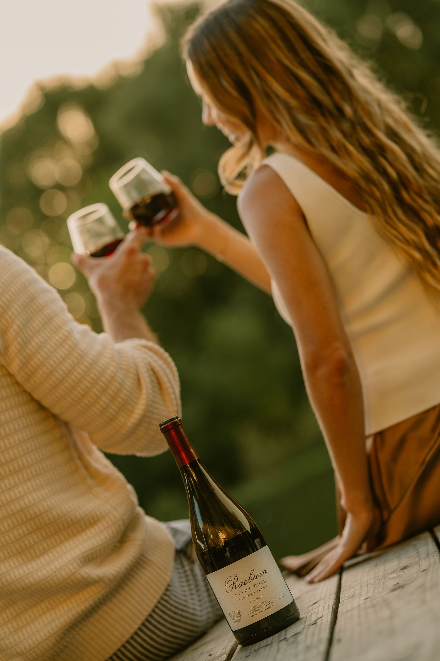 Couple toasting with red wine glasses at golden hour with a Raeburn wine bottle on a wooden surface.