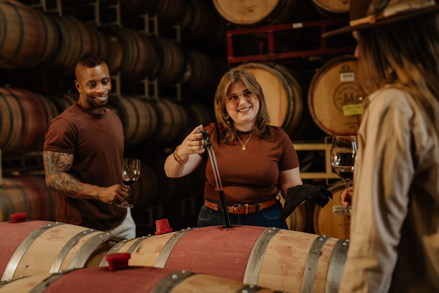 Three people enjoying a wine tasting tour inside a rustic winery barrel cellar.
