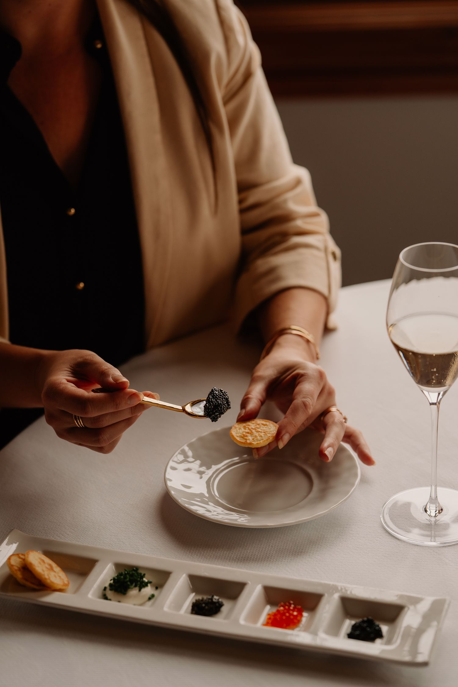 Hands serving caviar and accompaniments on white ceramic plates with champagne glasses in an elegant restaurant setting.