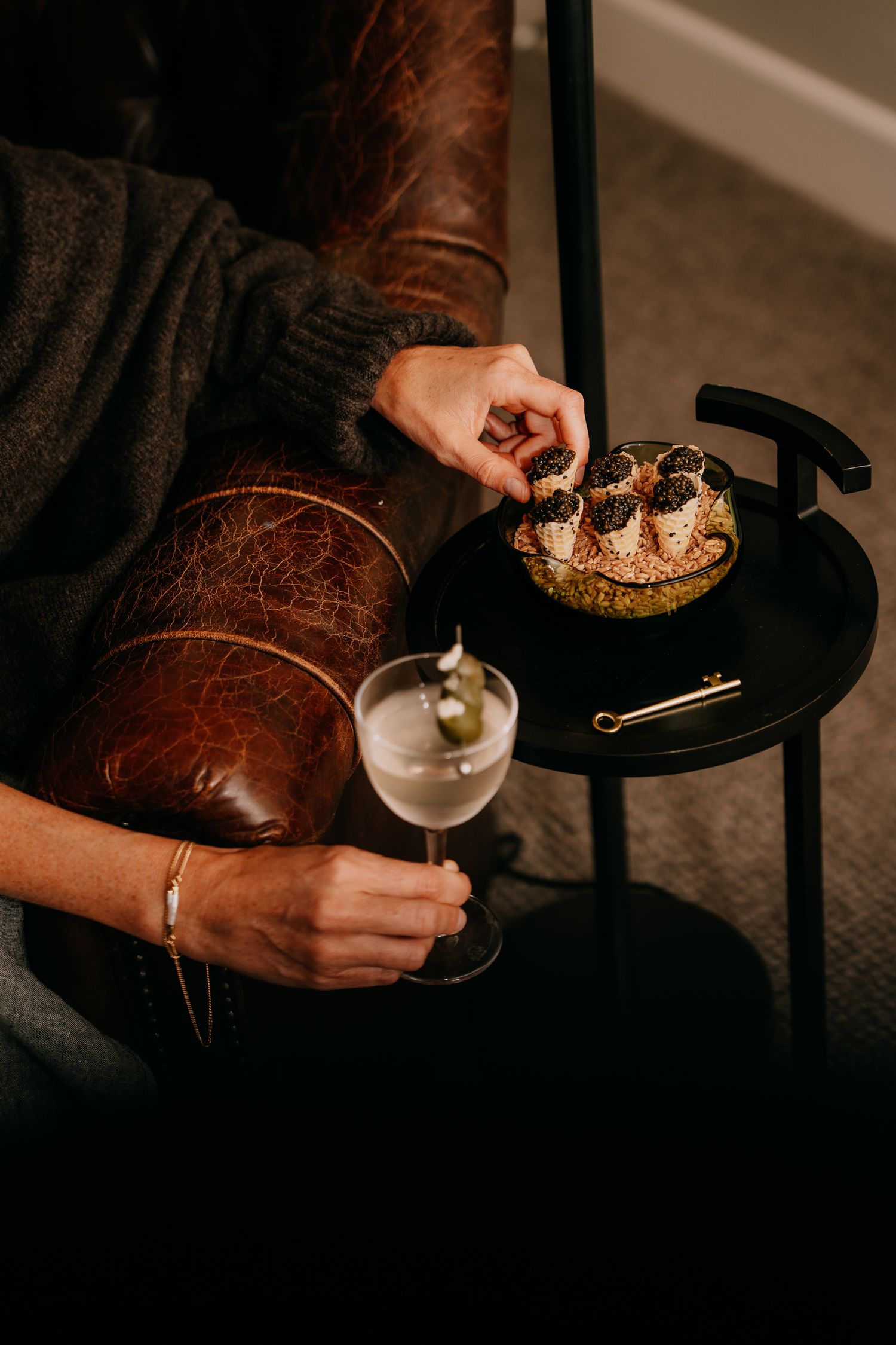 Someone in a brown leather jacket holds a martini glass while enjoying a decadent dessert on a small black side table.