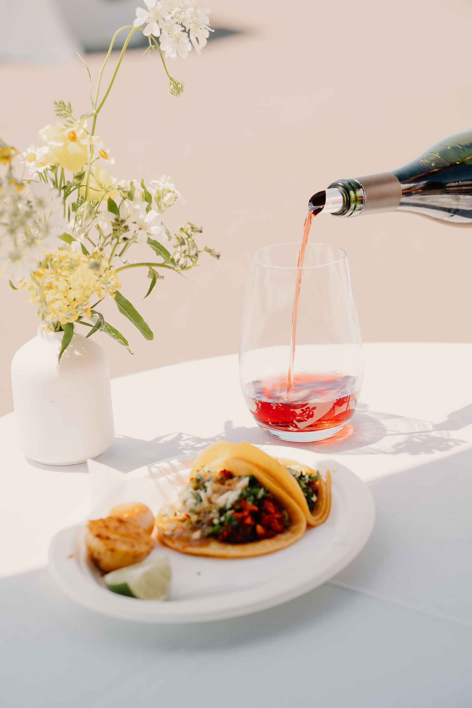 A table setting with rosé wine being poured into a glass alongside tacos and white flowers in a vase.