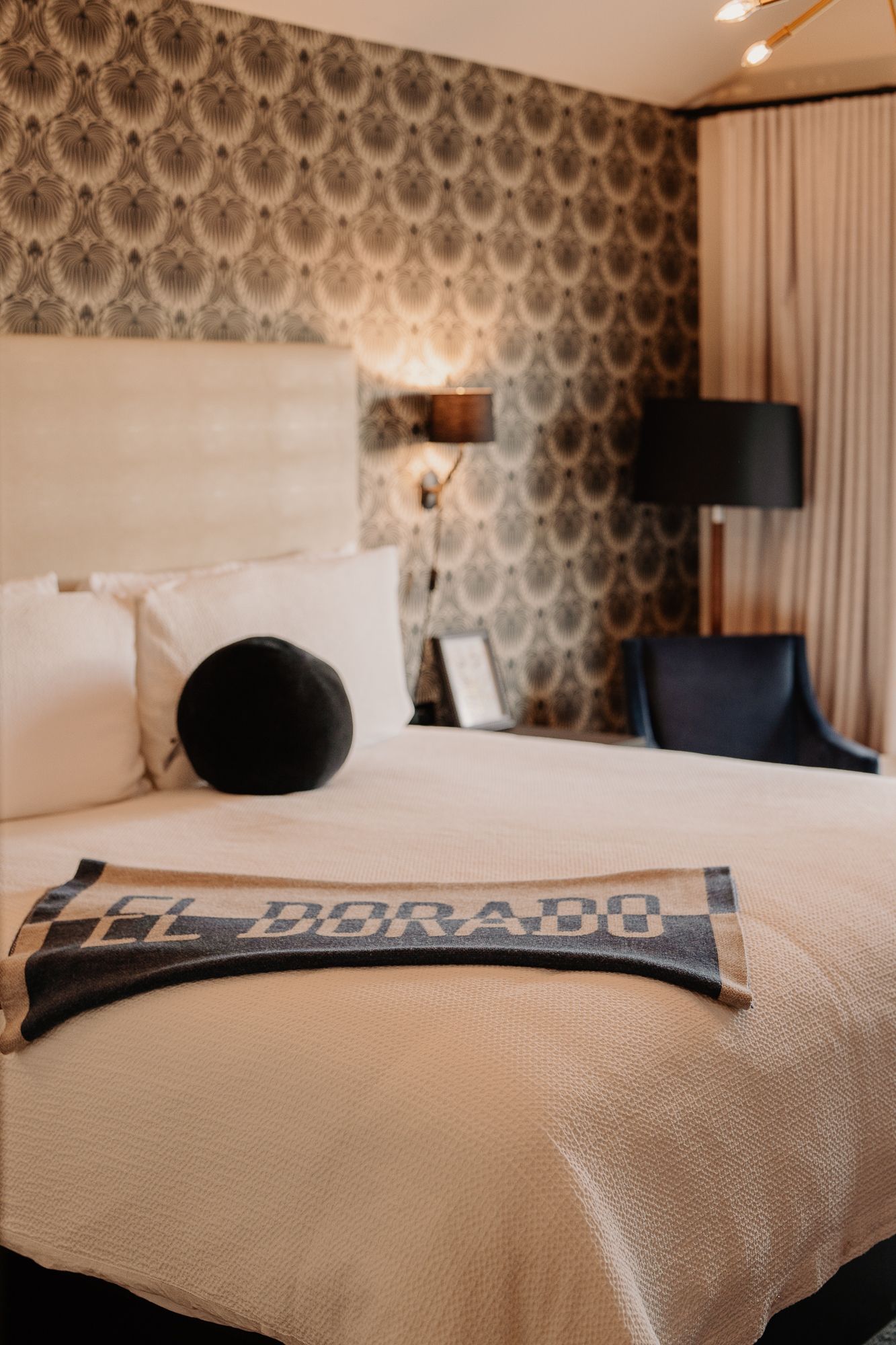 Modern hotel bedroom with damask wallpaper, white bedding, and black headboard against patterned wall.