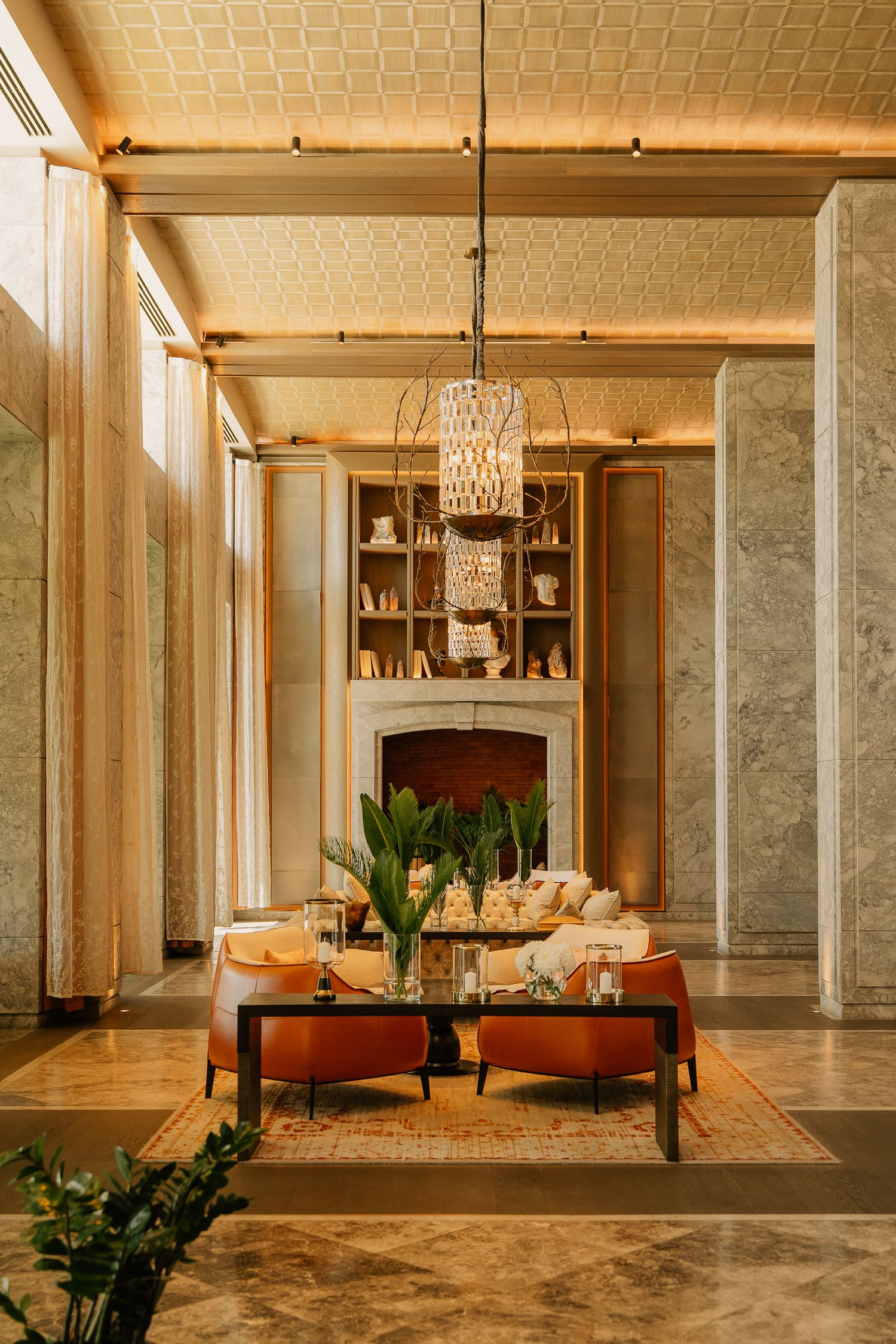 Elegant hotel lobby with crystal chandelier, marble columns, orange armchairs, and a fireplace with built-in bookshelves.
