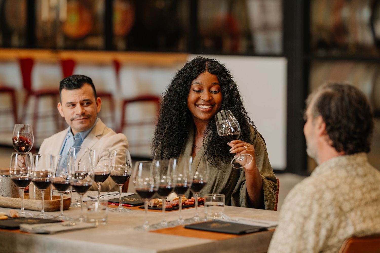 Guests smile and taste red wine at an elegant winery table set with multiple wine glasses and food pairings.
