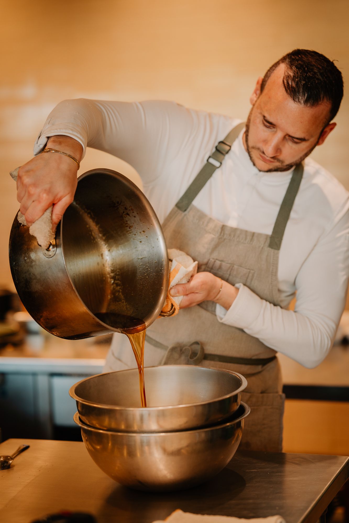 A chef in an apron pouring sauce from a pan into a stainless steel mixing bowl in a professional kitchen.