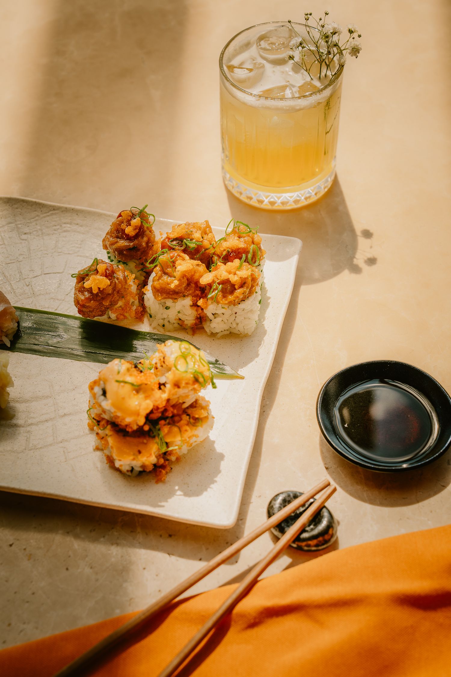Crispy tempura sushi rolls on white plate with chopsticks, soy sauce dish, and iced cocktail garnished with flowers.