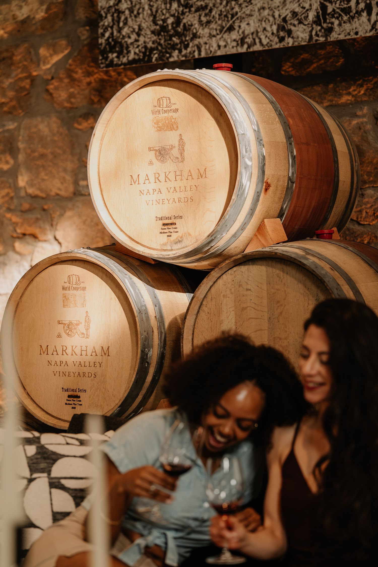Two women enjoying red wine beside Markham Napa Valley Vineyards oak barrels.