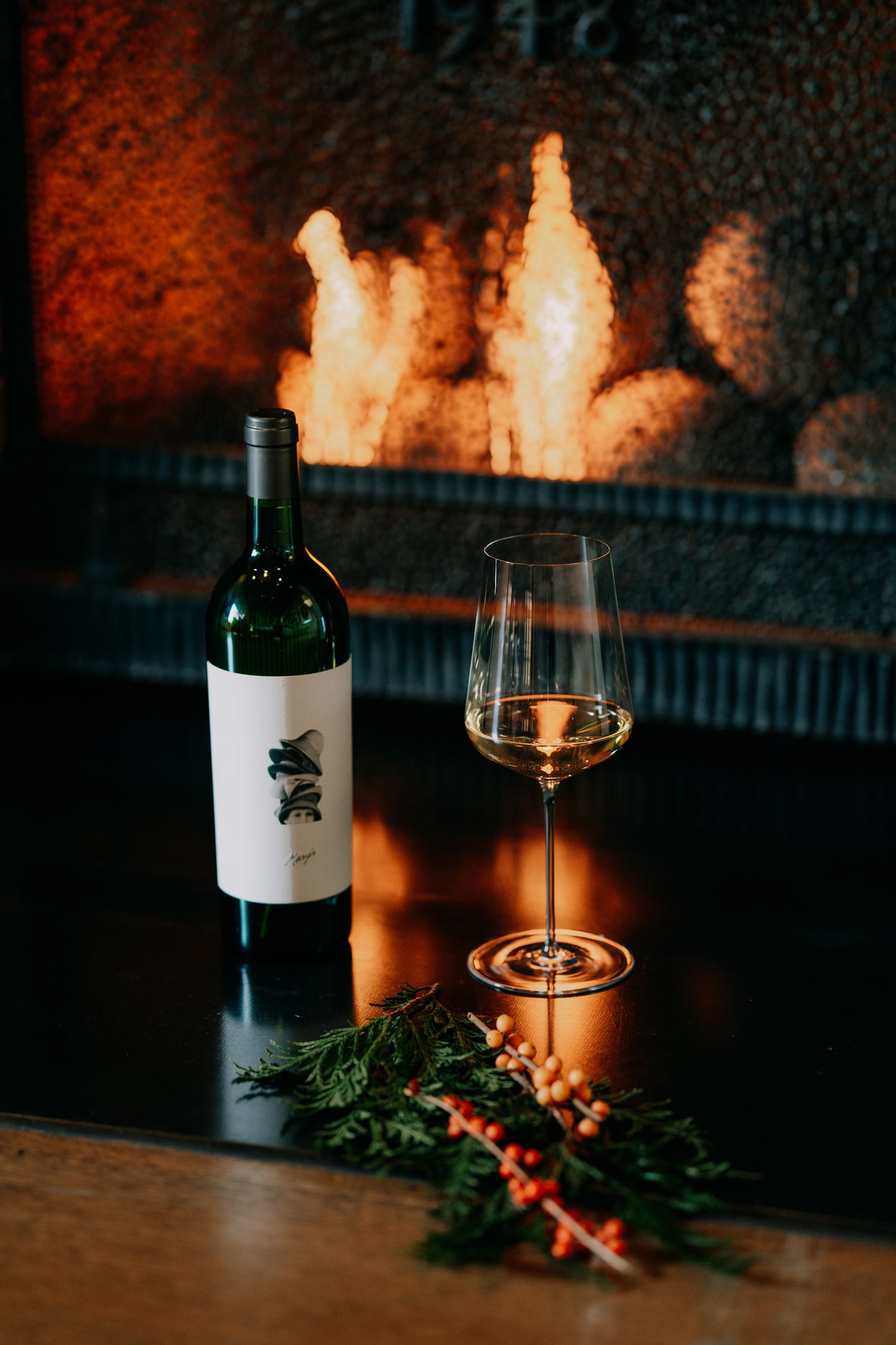 Wine bottle and glass with holiday greenery and berries on dark table with warm fireplace glow in background.