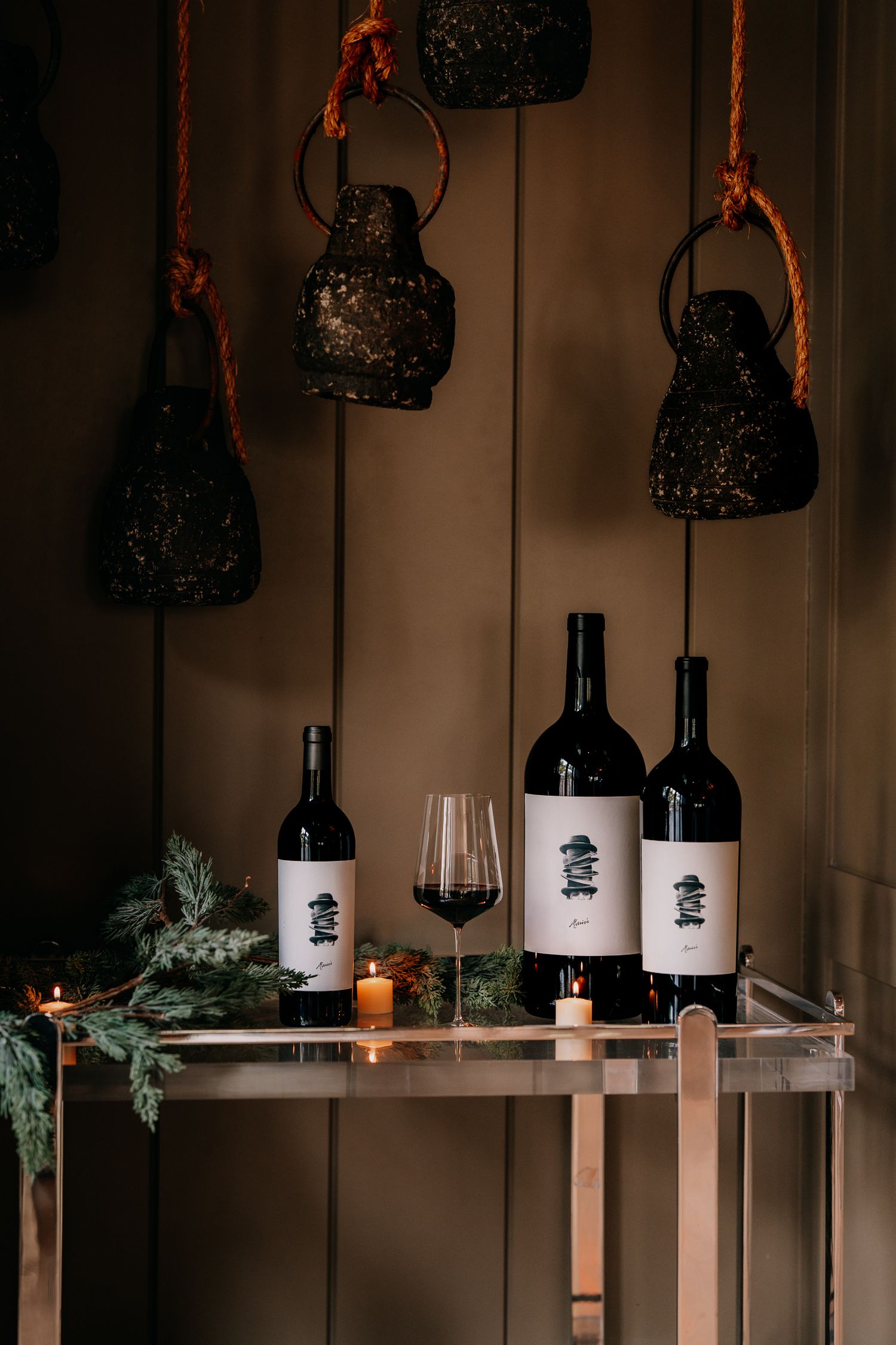 Dark wine bottles with minimalist black and white labels displayed on wooden shelves with hanging vintage teapots above.