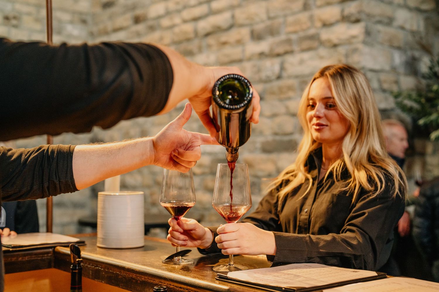 A sommelier pours red wine into a glass for a smiling blonde woman at an elegant stone-walled restaurant.