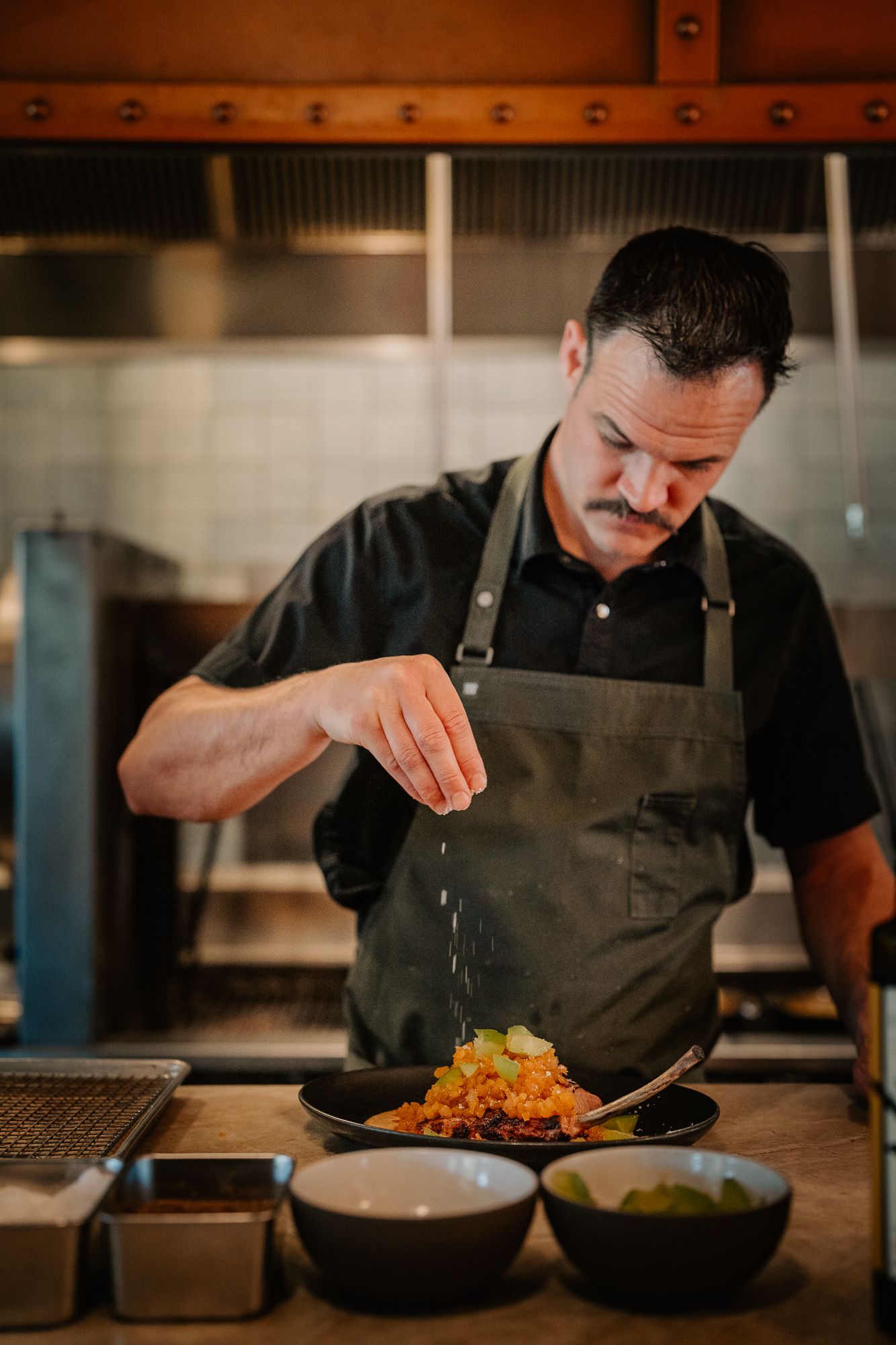 Chef in dark apron carefully seasoning a colorful dish in a professional restaurant kitchen.
