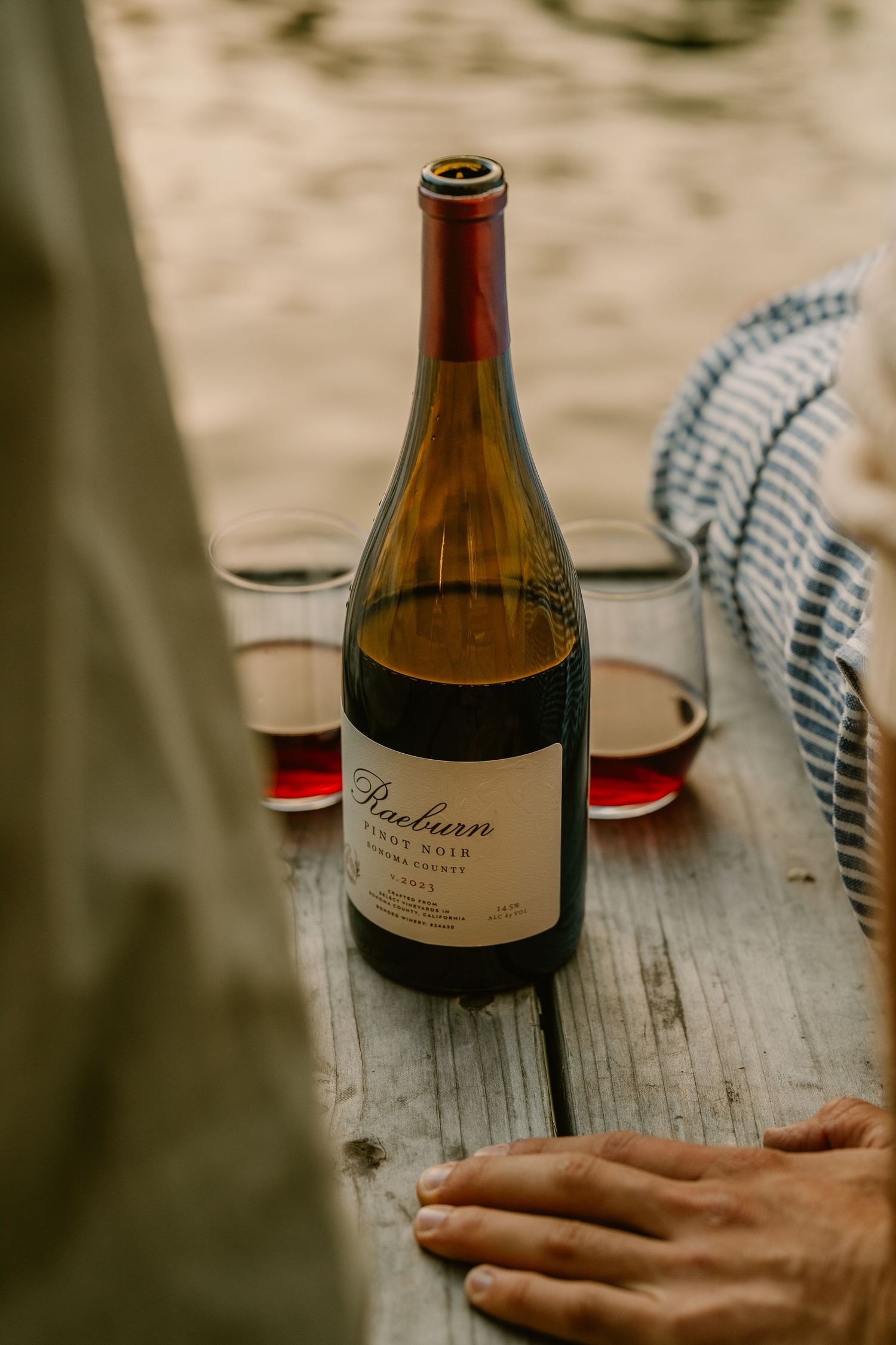 Raeburn Pinot Noir wine bottle on rustic wooden table with two glasses of red wine outdoors.