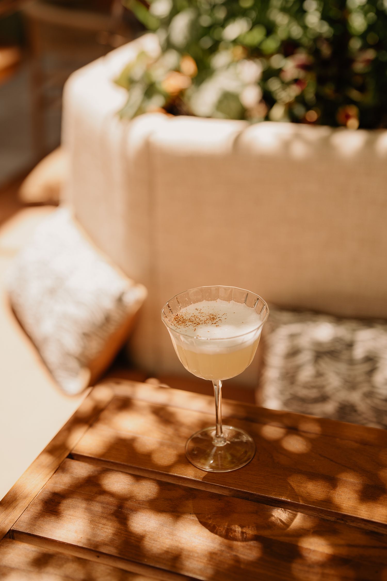 Sunlit cocktail glass on wooden table with dappled shadows and soft cushioned seating.
