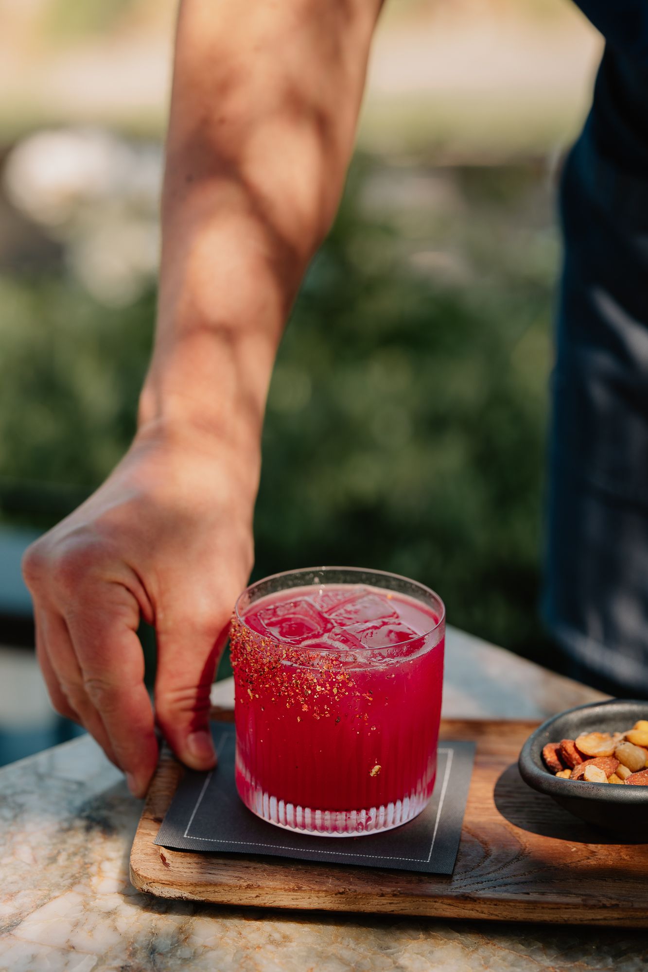 A bartender places a vibrant red cocktail on ice on a wooden serving board outdoors.