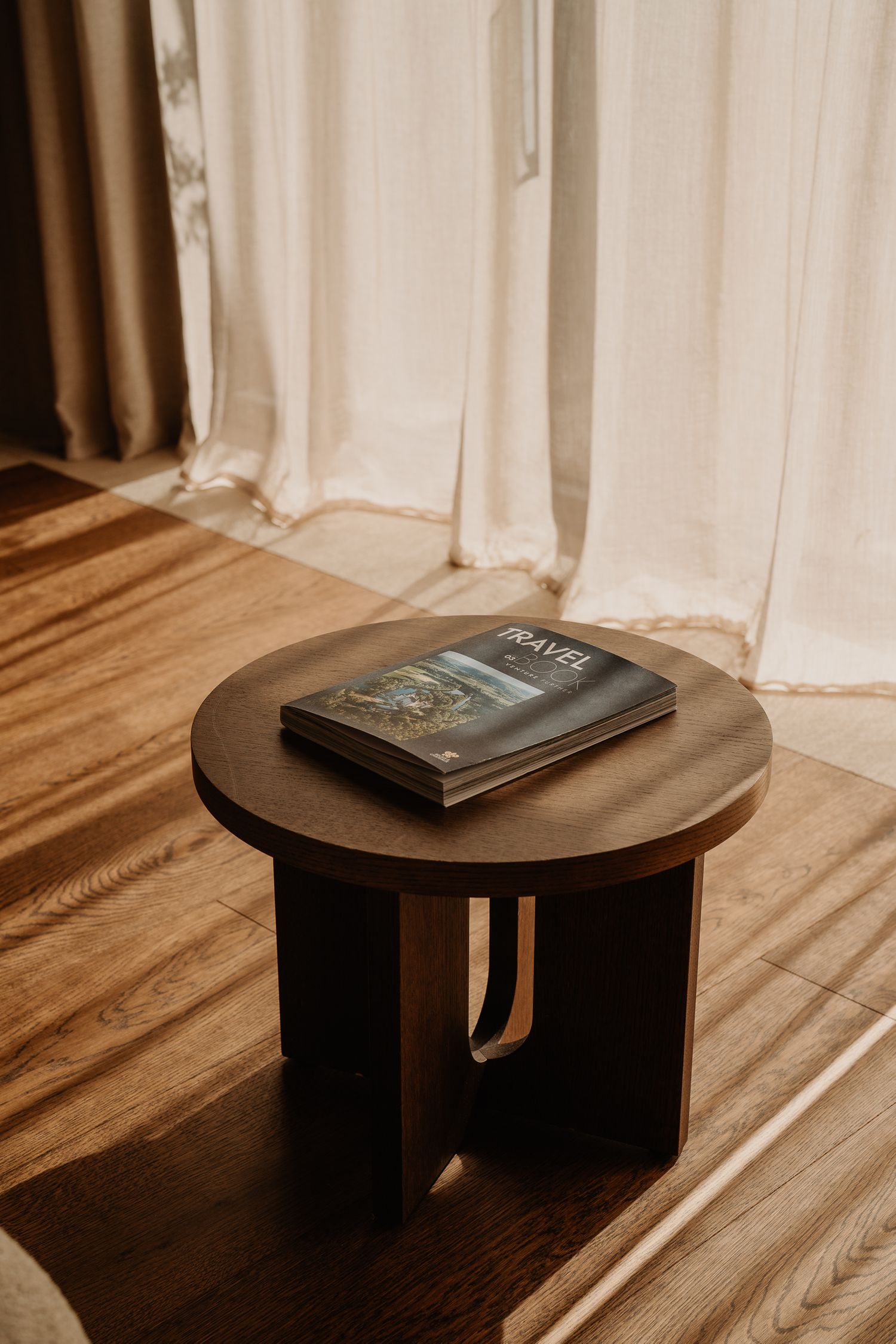 A dark book rests on a round wooden side table with geometric legs against white curtains and hardwood flooring.