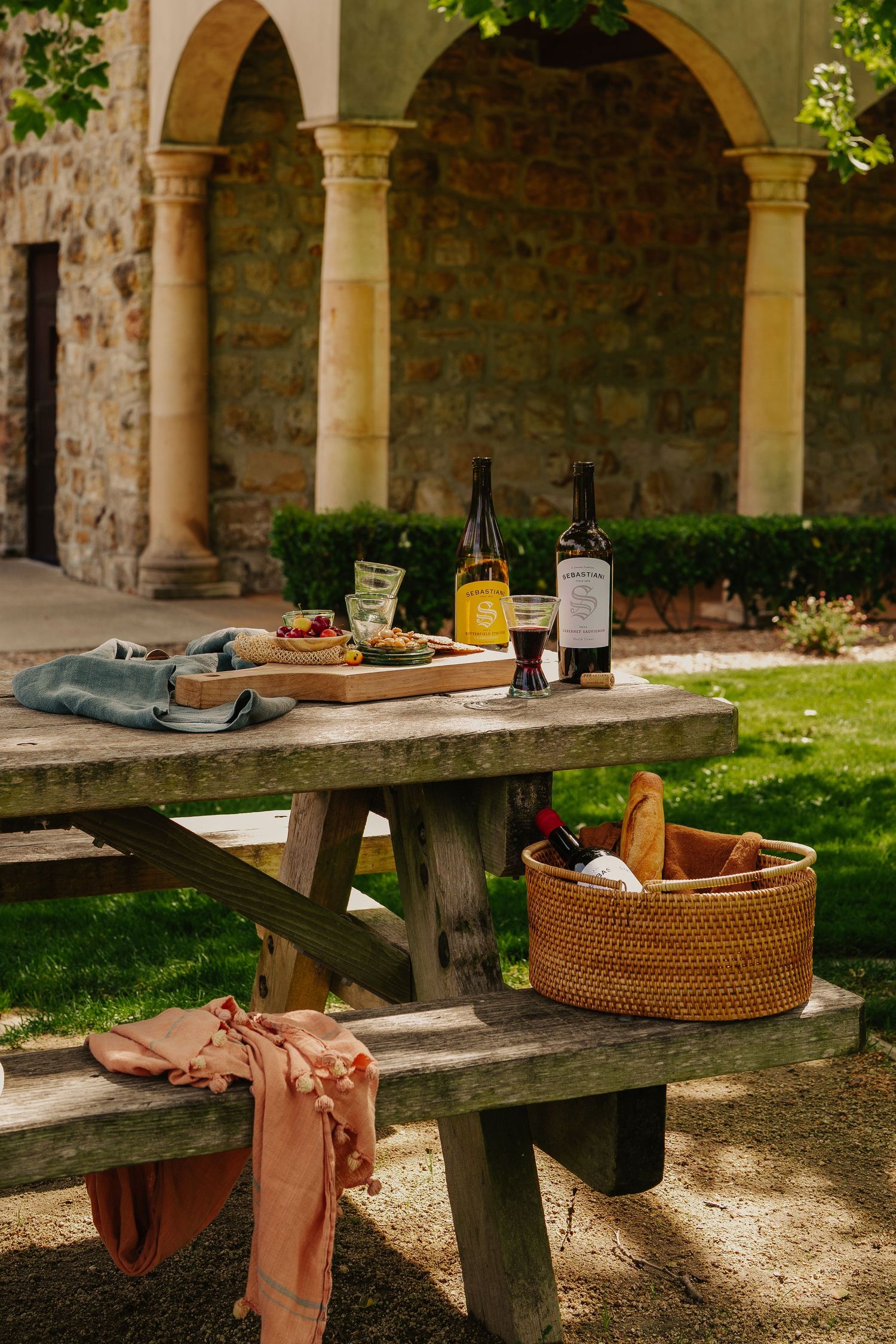 Rustic picnic table with Sebastiani wine bottles, food spread and wicker basket in front of stone arches.