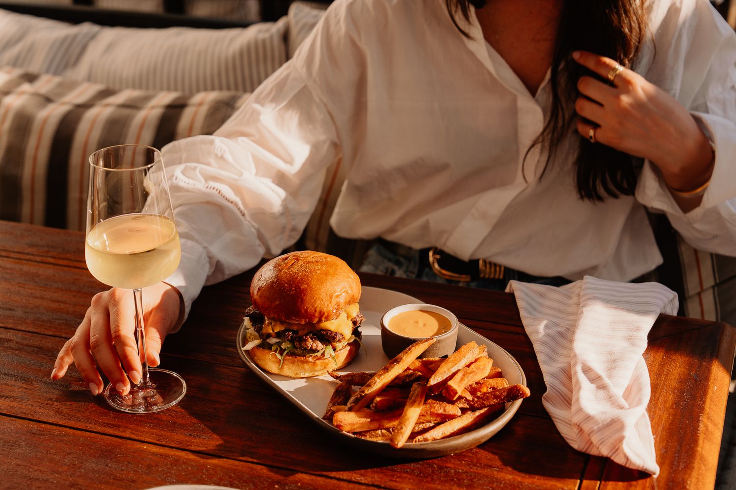 Woman in white shirt enjoying a gourmet burger with fries and white wine at a wooden table.