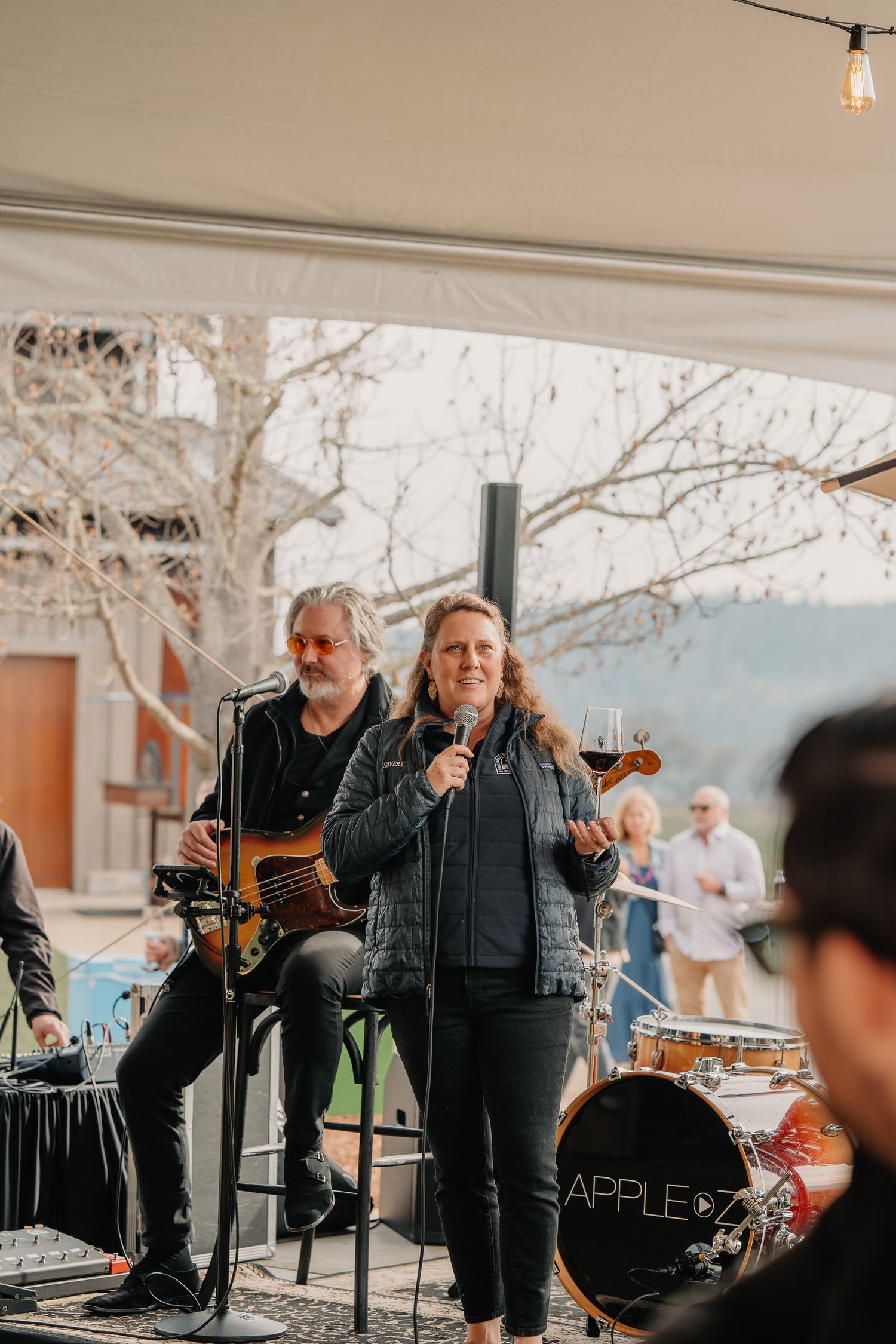 Woman speaks into microphone at Silver Oak Winery event with live band performing in background.