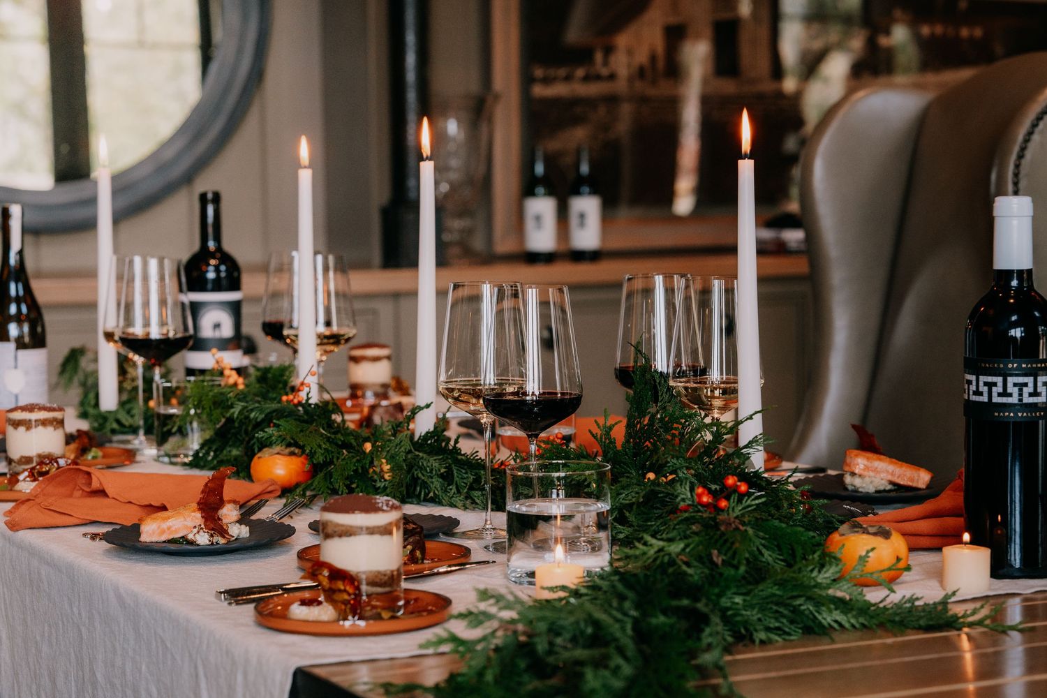 Elegant table setting with candles, wine glasses, and evergreen branches decorating a rustic wooden dining table.