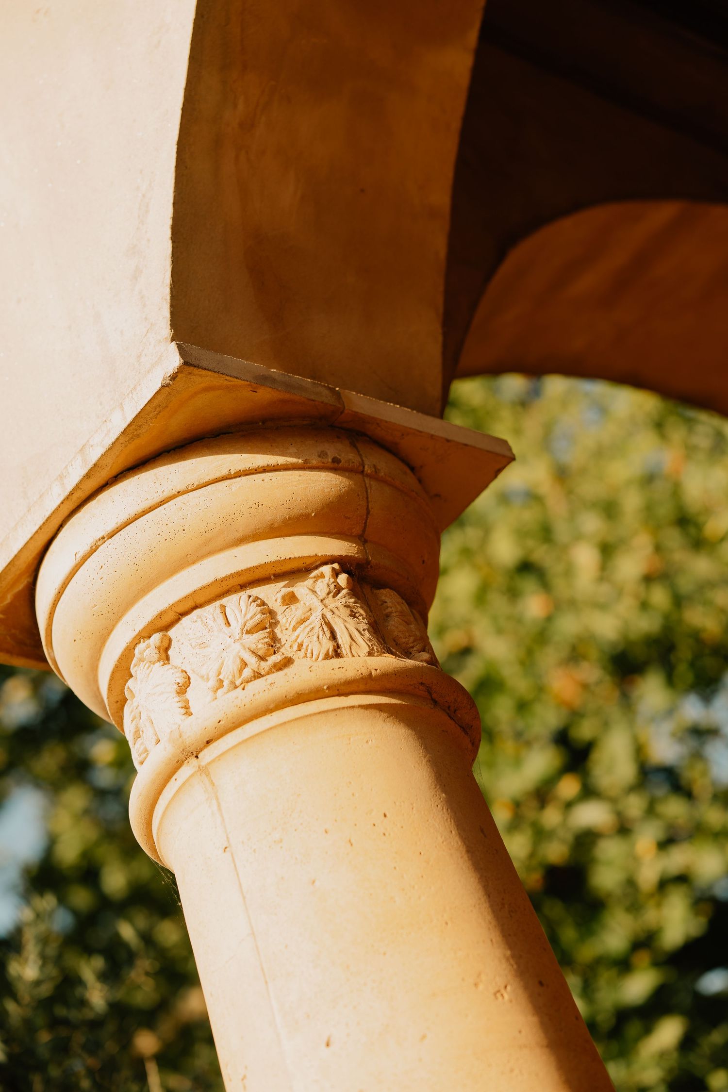 Close-up of an ornate stone column capital with carved floral details supporting a terracotta roof structure.