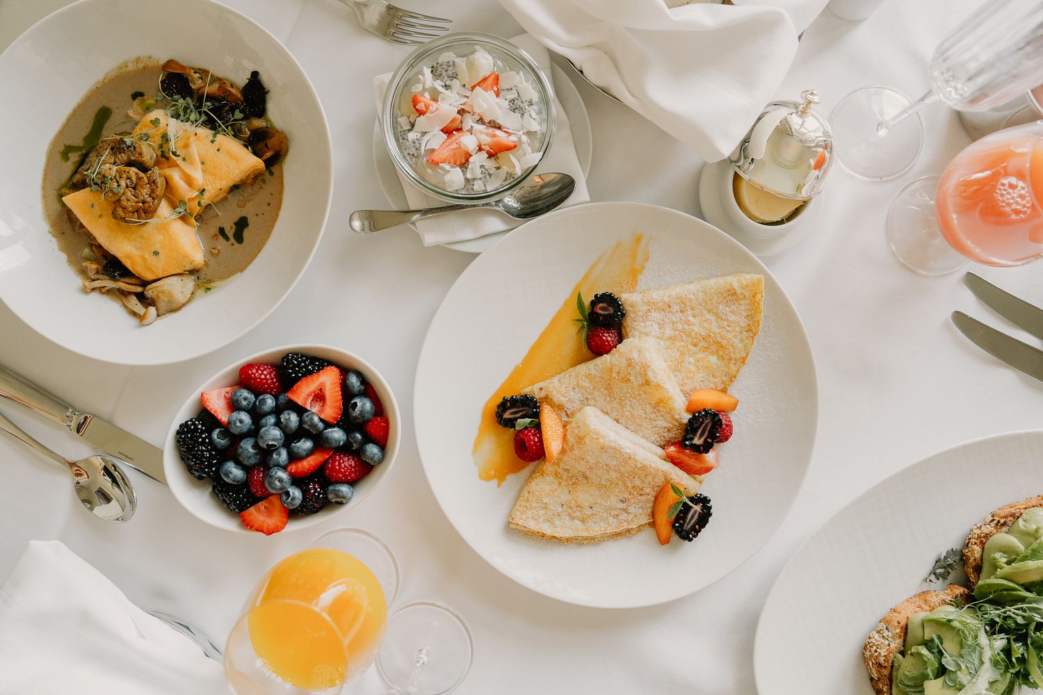 Elegant brunch spread with crepes, fresh berries, orange juice, and salads on a white tablecloth.